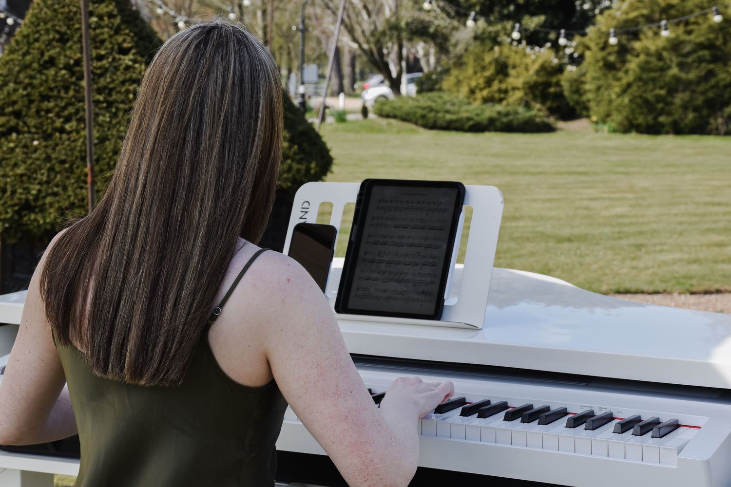 Young woman playing a white grand piano outdoors using a tablet for sheet music.