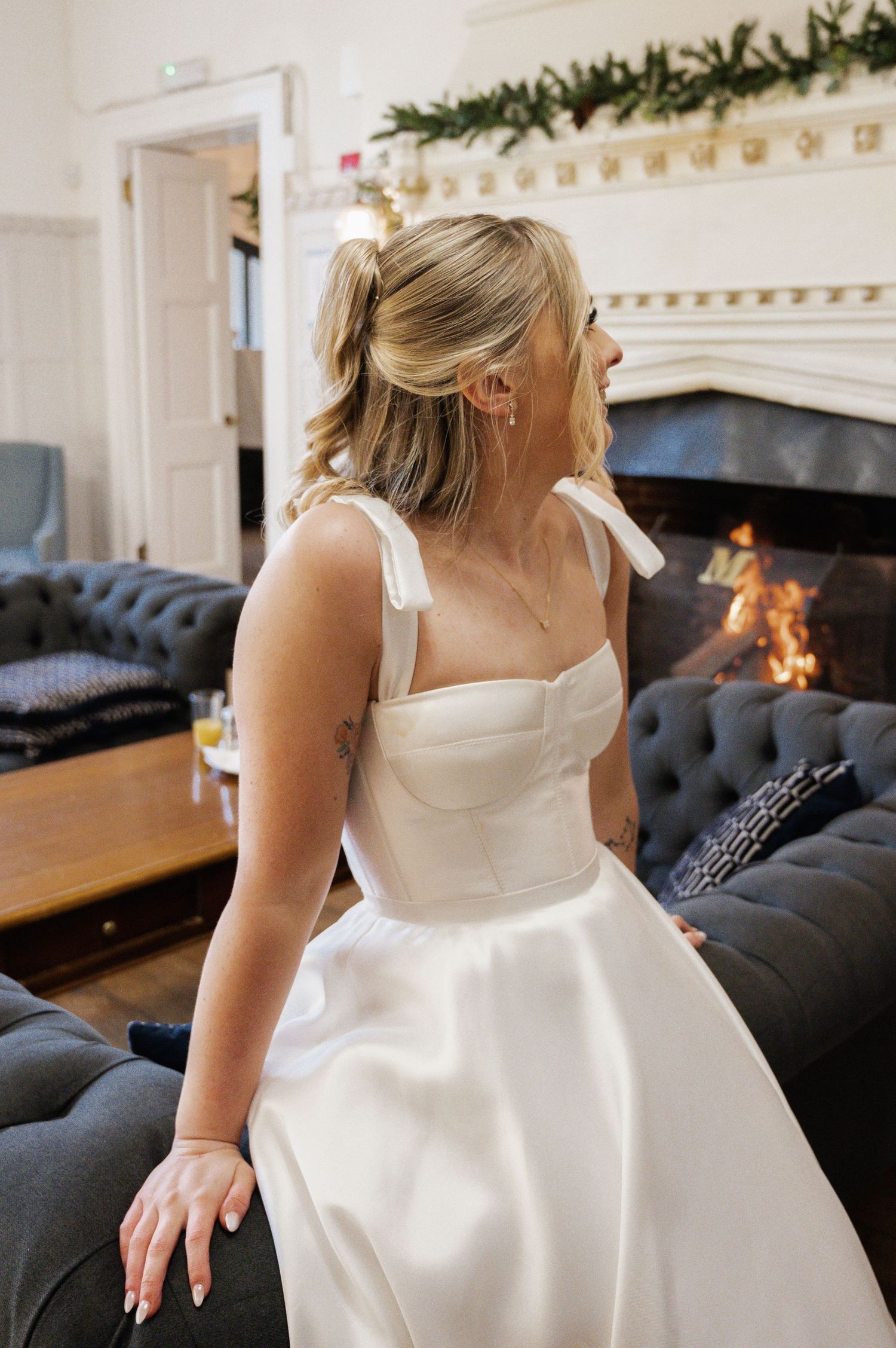 Bride in elegant white satin corset wedding dress seated by a roaring fireplace in a luxury venue.