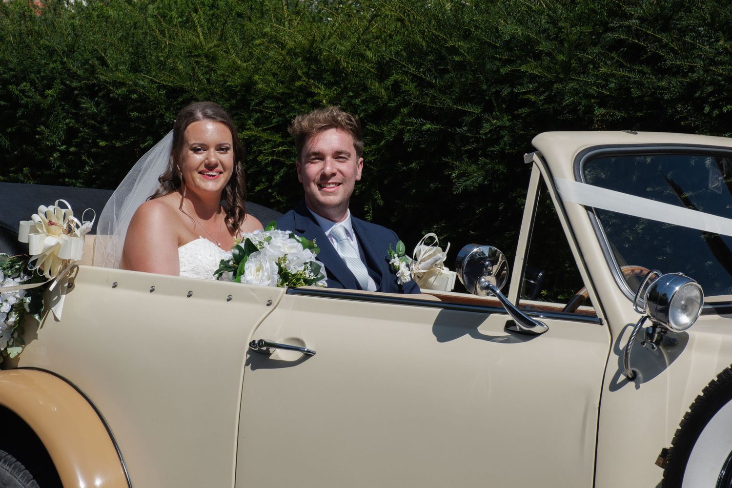 Smiling bride and groom pose in a classic vintage cream wedding car decorated with white floral arrangements.