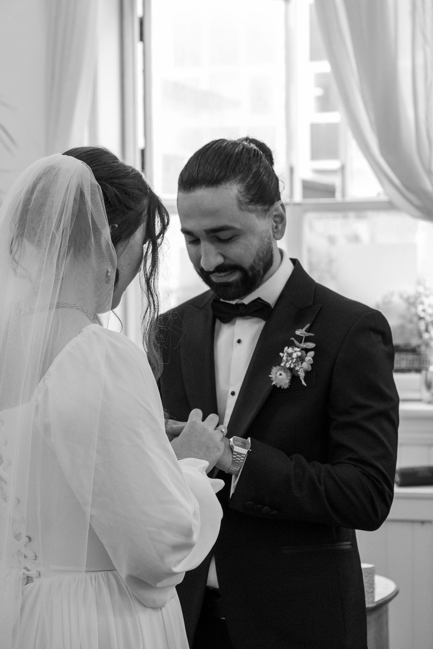 Black and white photo of groom smiling warmly while exchanging rings with bride during an intimate wedding ceremony.