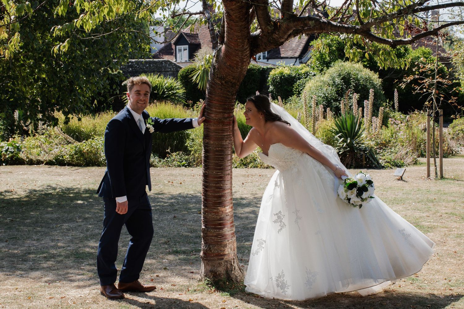 Playful bride and groom sharing lighthearted moment around tree trunk in sunny garden setting.