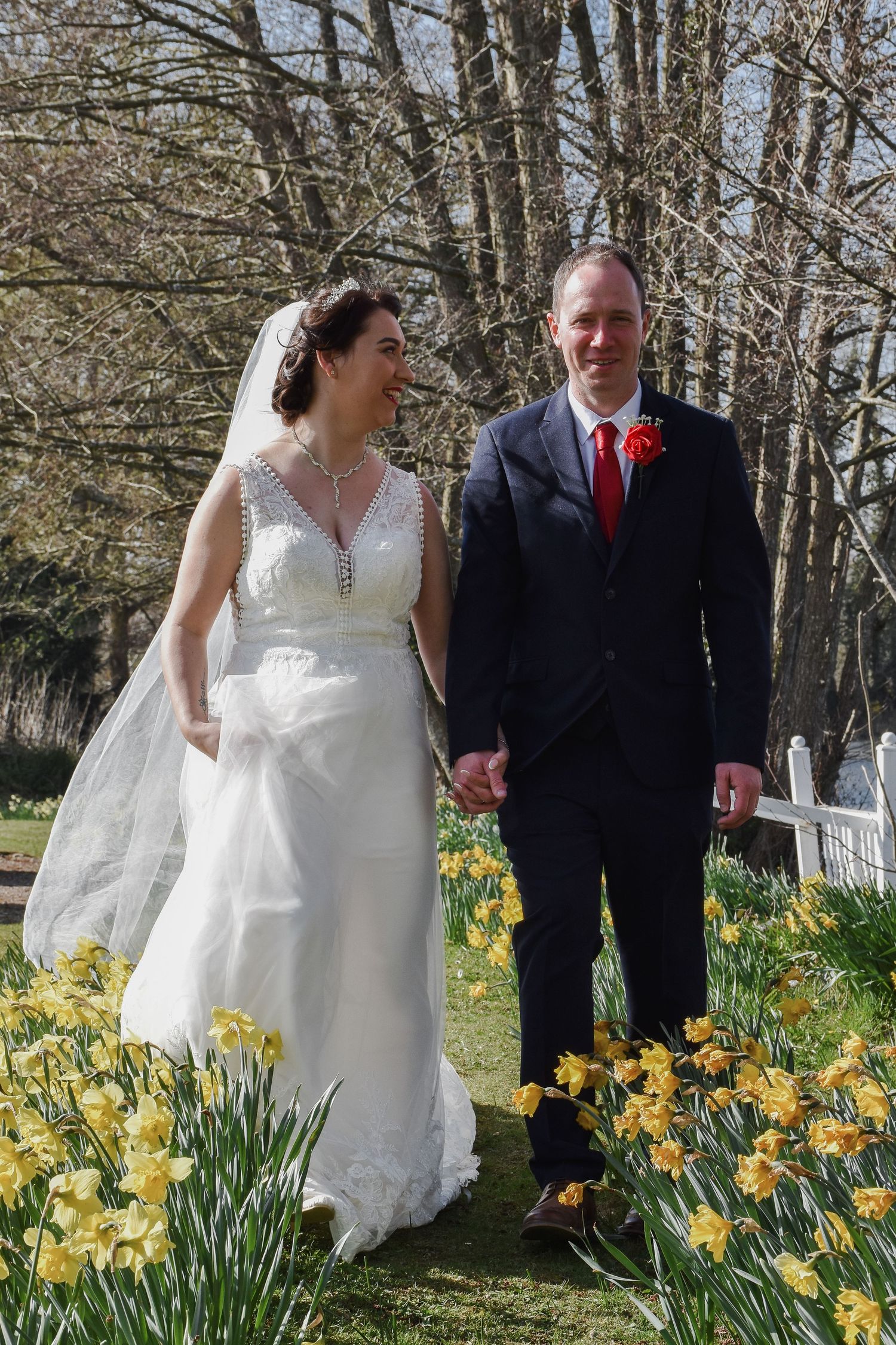 Smiling bride and groom walking hand in hand through a field of yellow daffodils on their spring wedding day.