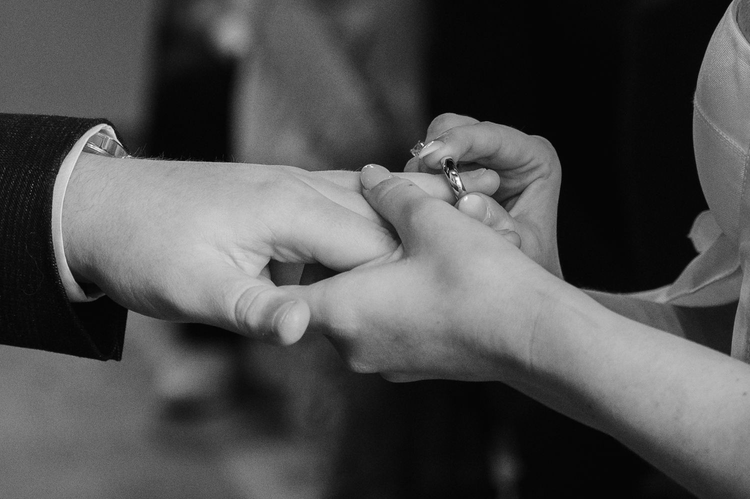 Black and white close-up of groom placing wedding ring on brides finger during wedding ceremony.