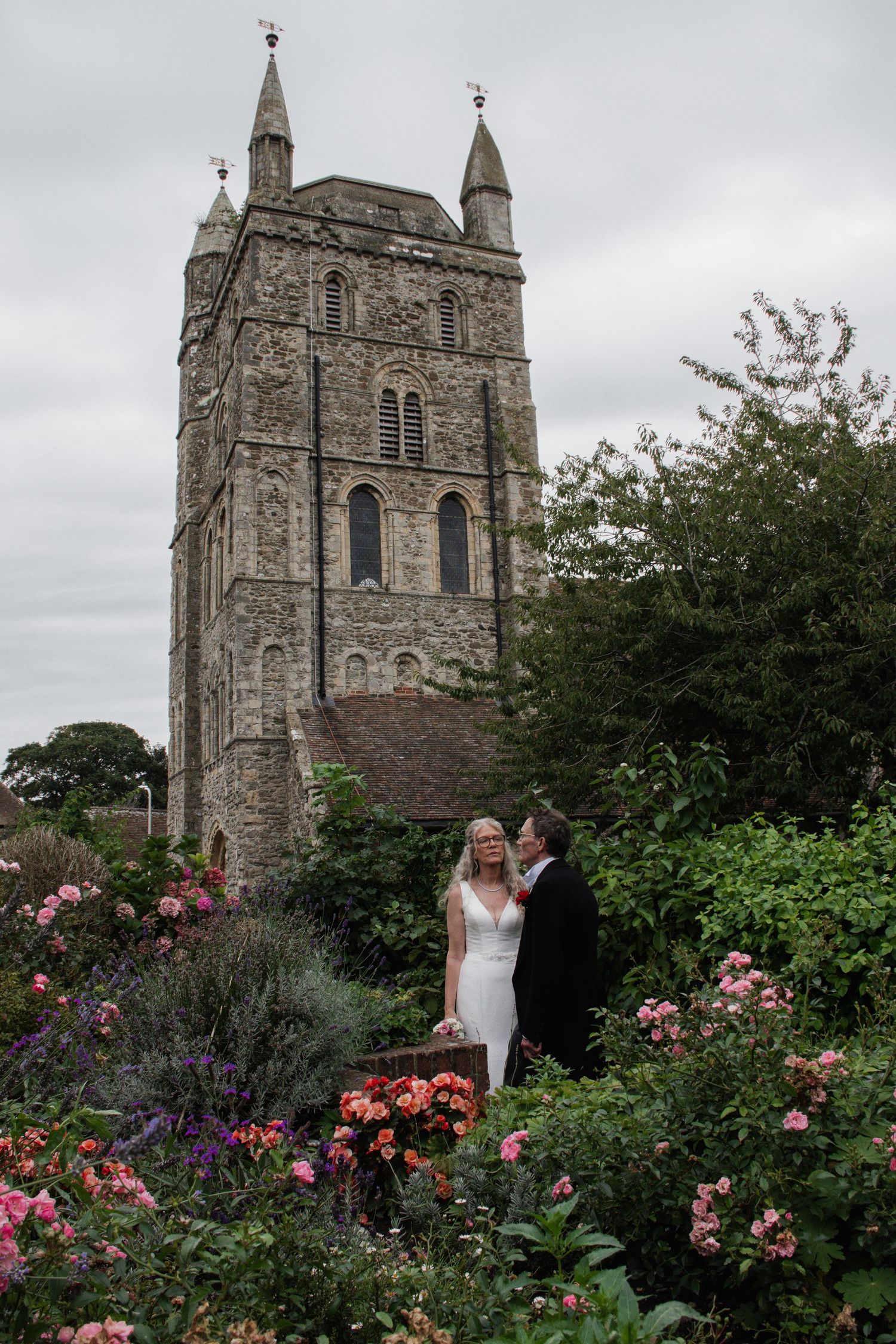 Bride and groom stand romantically among colorful roses in a lush garden with a historic stone church tower behind them.