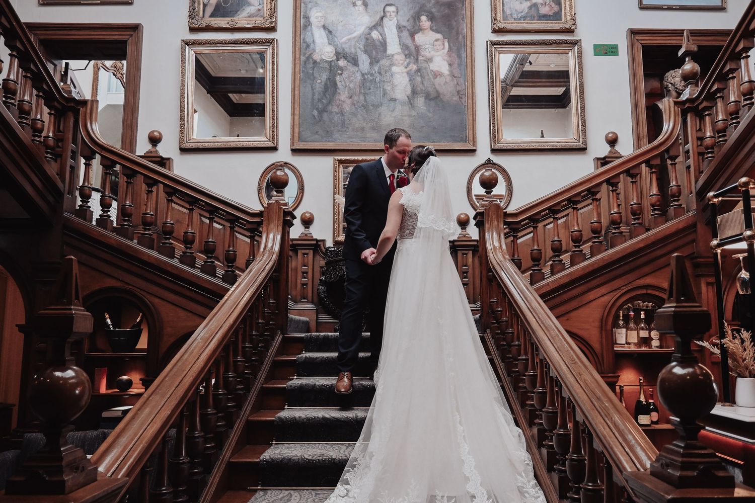 Bride and groom sharing a kiss on an ornate wooden staircase inside a grand historic mansion.