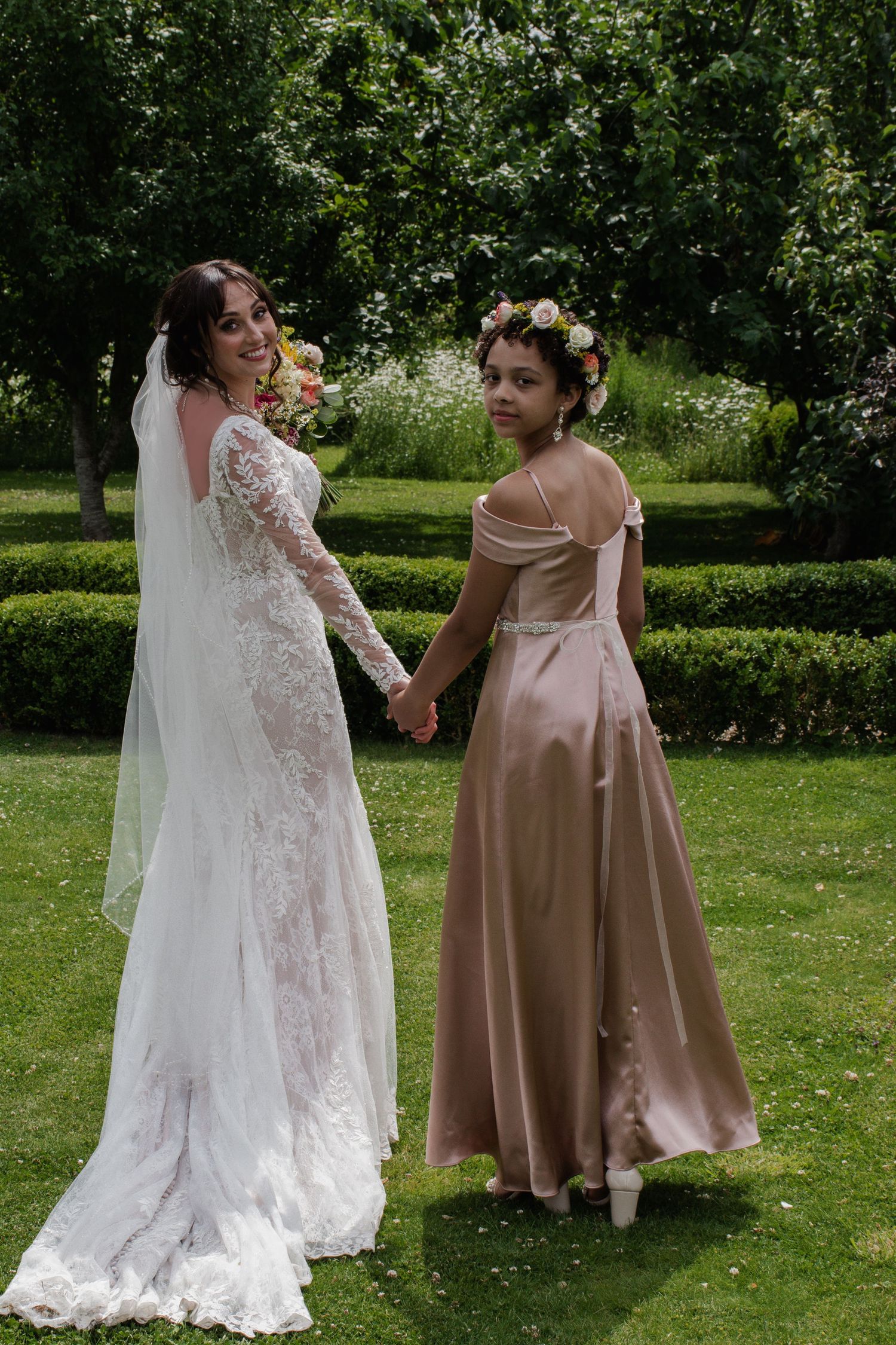 Bride in lace gown holds hands with young bridesmaid in champagne dress in garden setting.