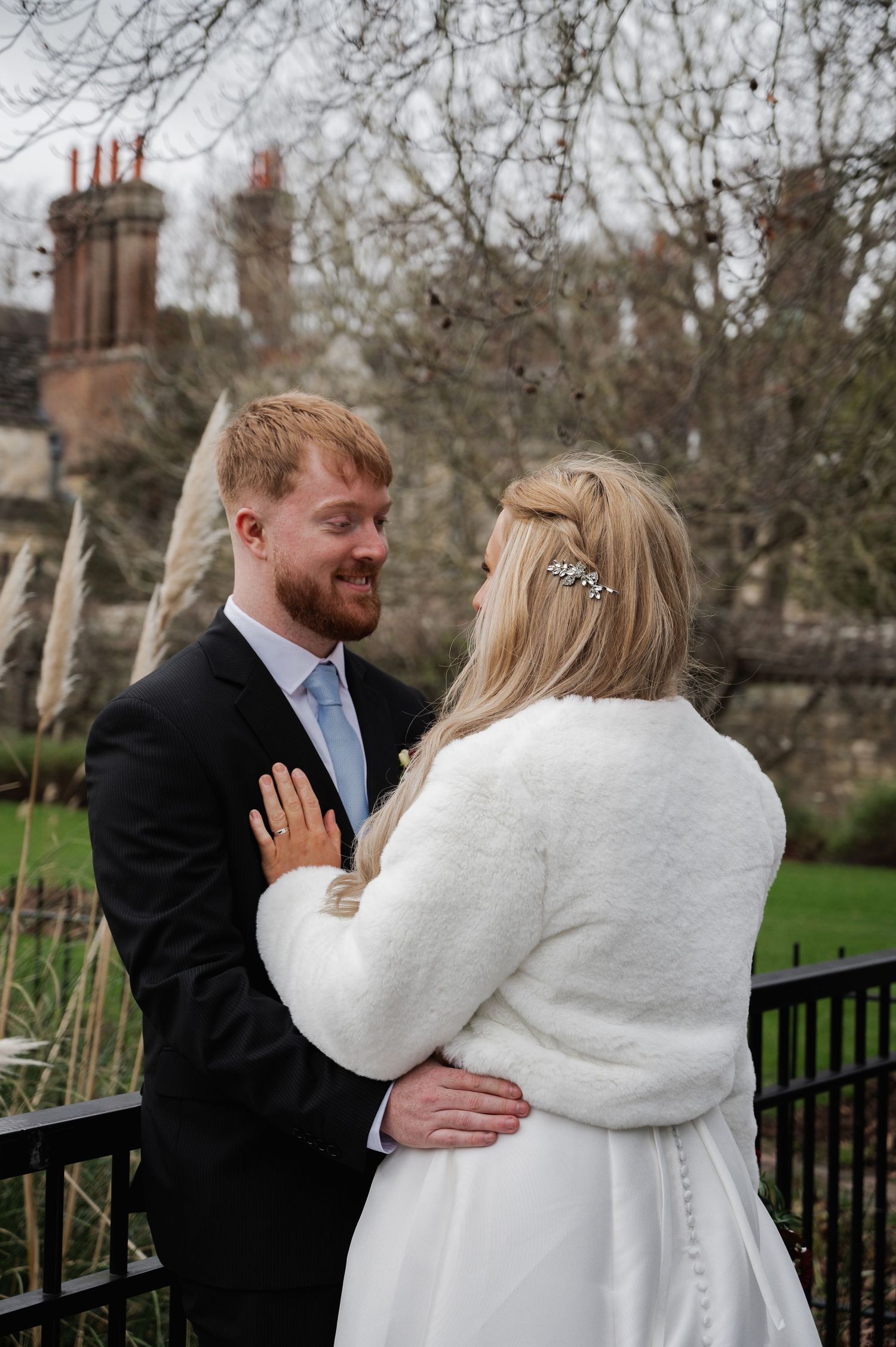 Smiling groom in black suit embraces bride in white fur coat at outdoor winter wedding venue.