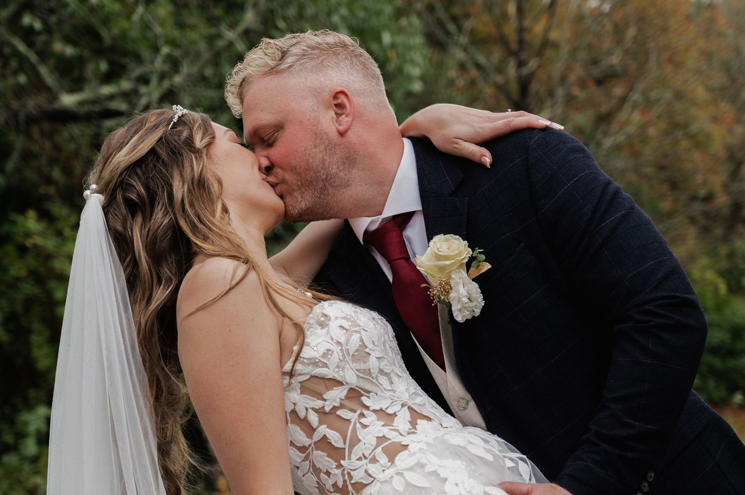 Bride and groom sharing a passionate kiss outdoors with autumn trees in the background.