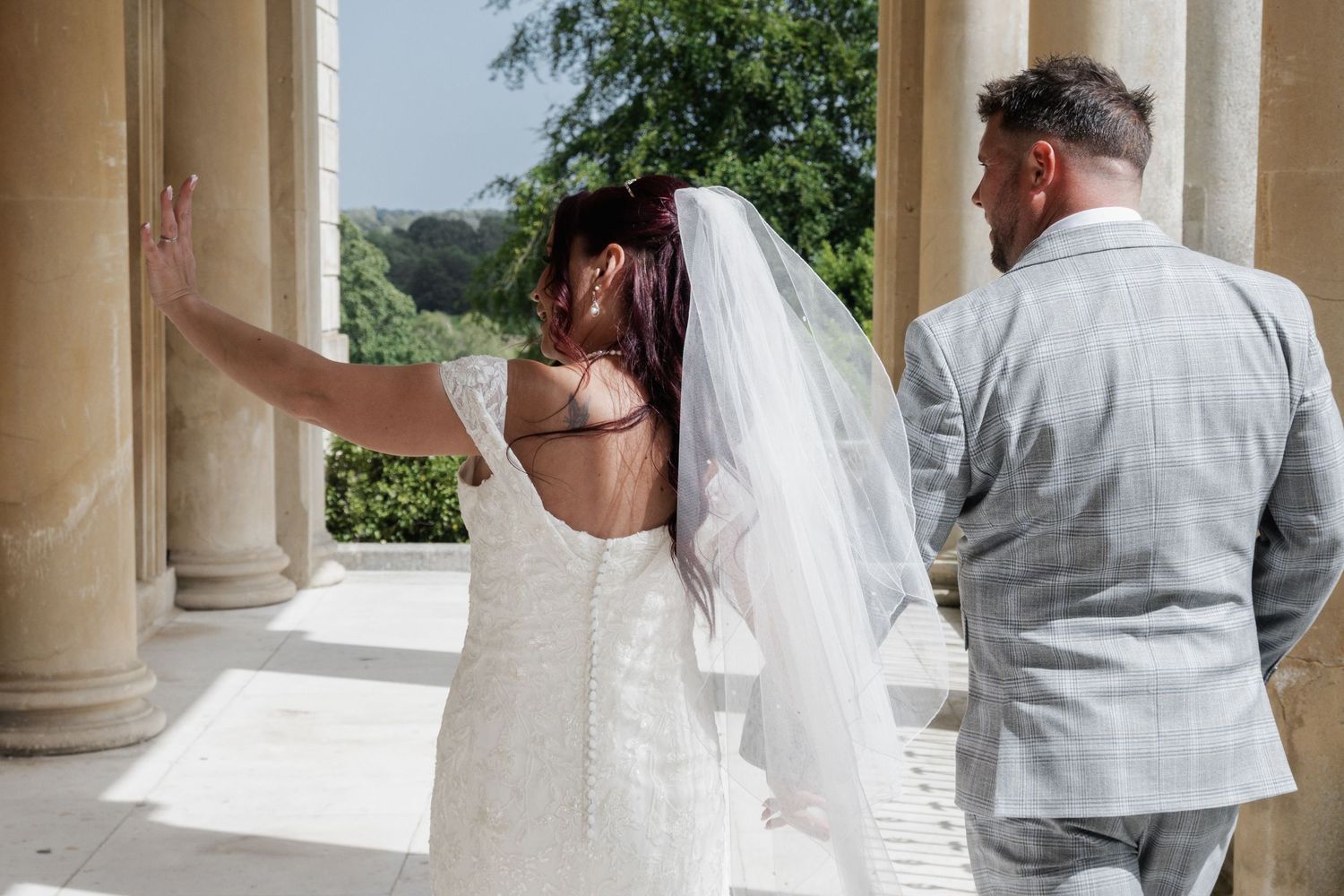 A bride in a backless lace gown and groom walk between grand columns at their wedding venue.
