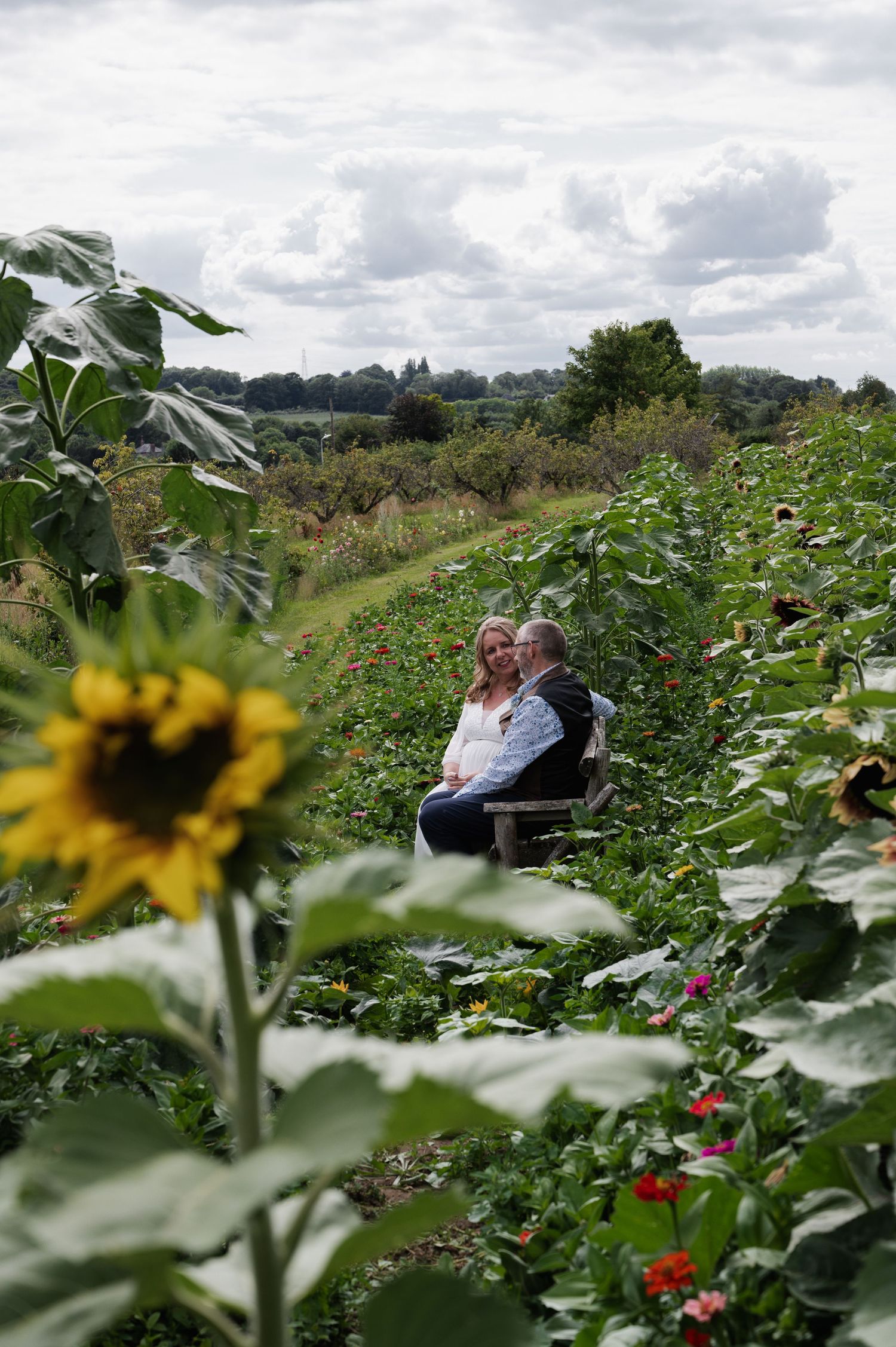 Bride and groom sitting among sunflowers and wildflowers in lush garden setting.