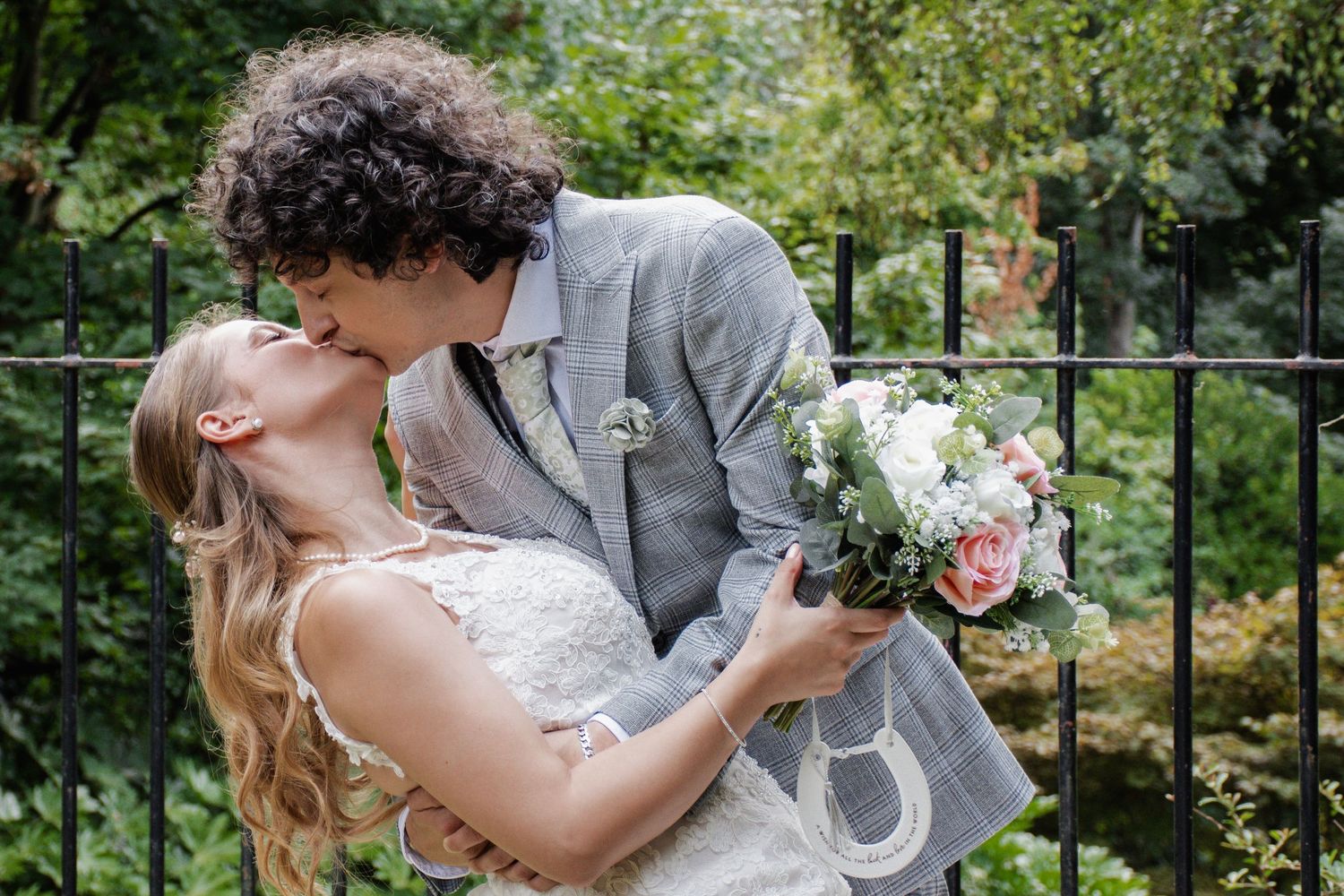 A groom dips his bride for a kiss as she holds a bouquet of pink and white roses outdoors.