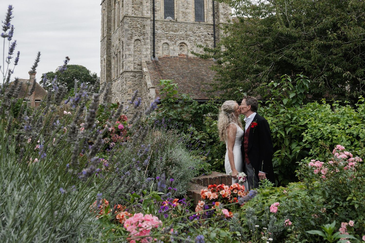 Bride and groom share a tender kiss among vibrant lavender and roses in a garden beside a historic stone church.