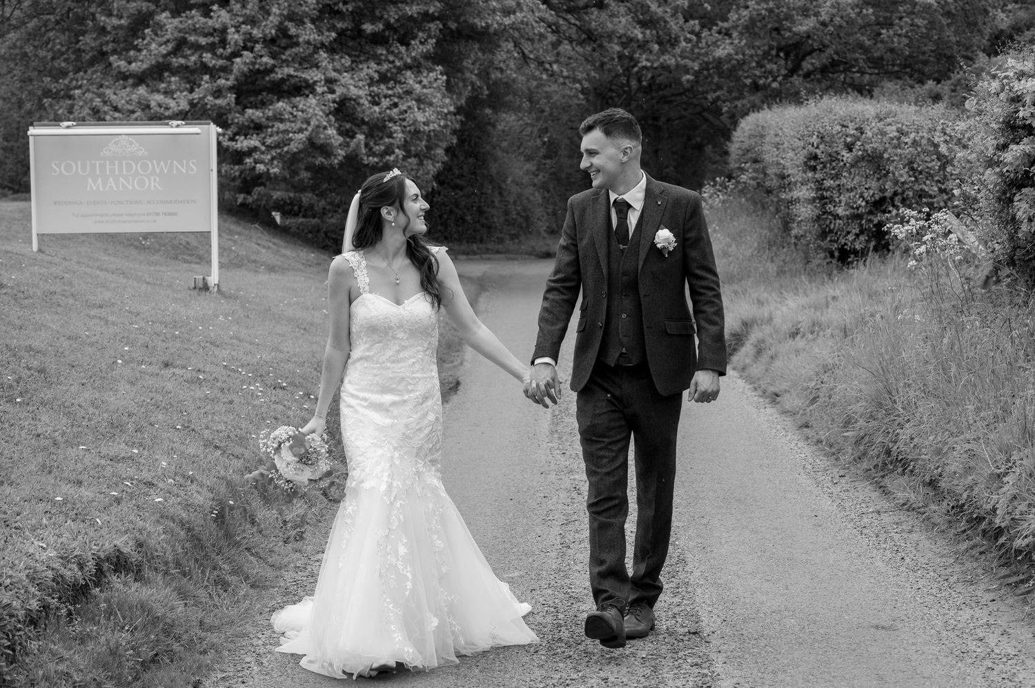 Black and white photo of smiling bride and groom walking hand in hand along a country path. Southdowns Manor