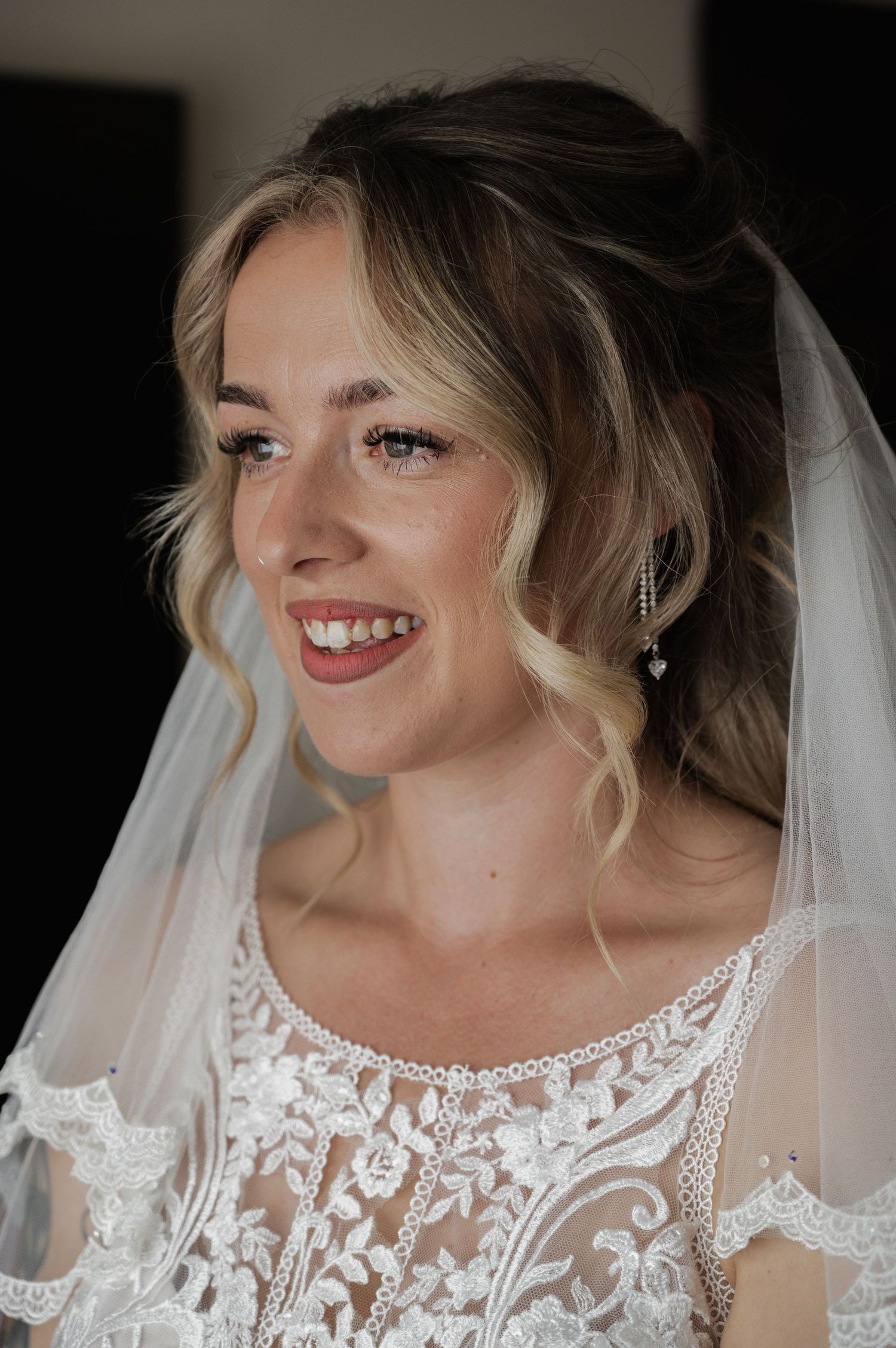A smiling blonde bride wearing a lace wedding dress and veil with delicate drop earrings on her wedding day.