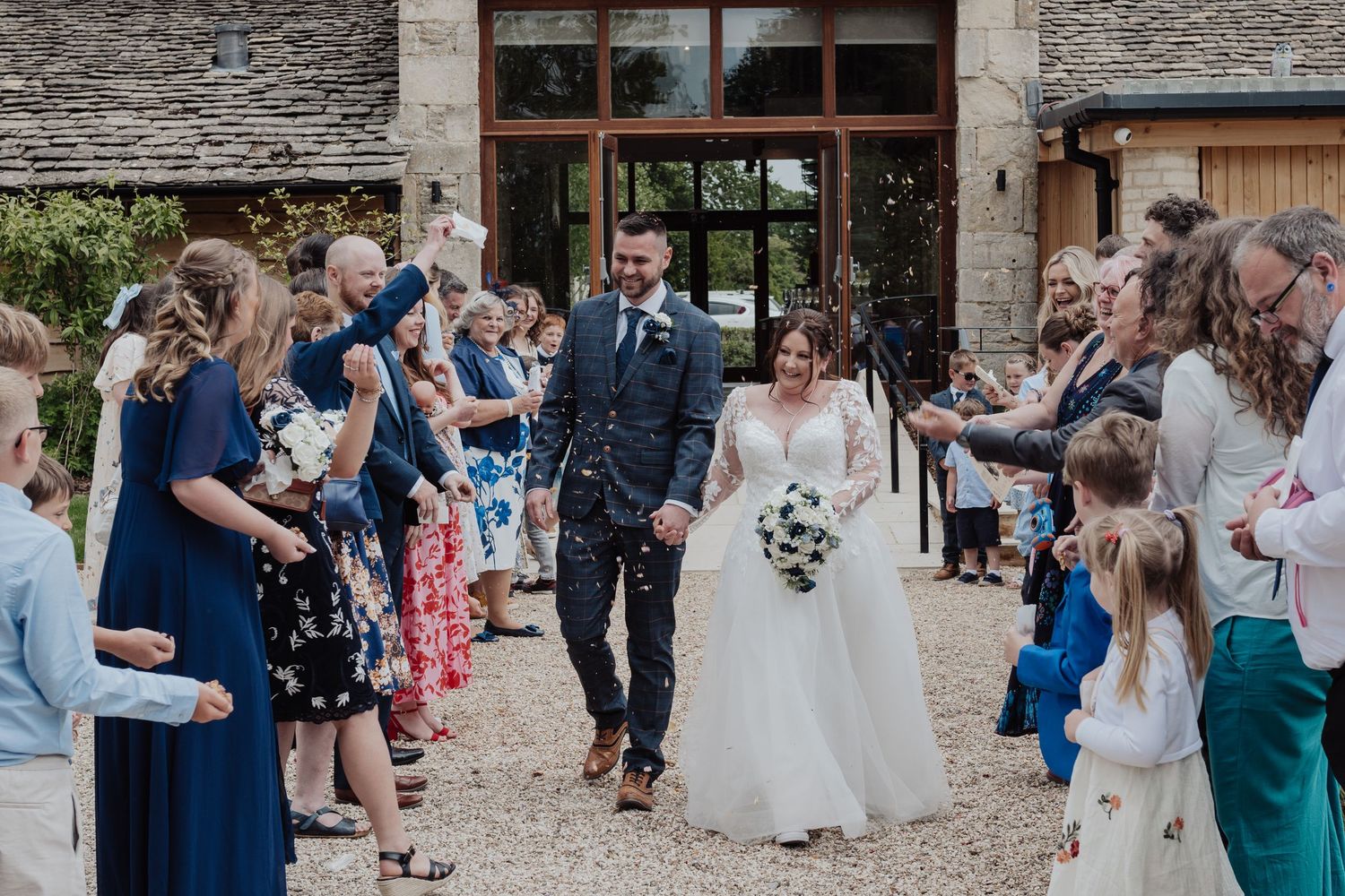Newlyweds walking through confetti shower with cheering guests outside barn venue.