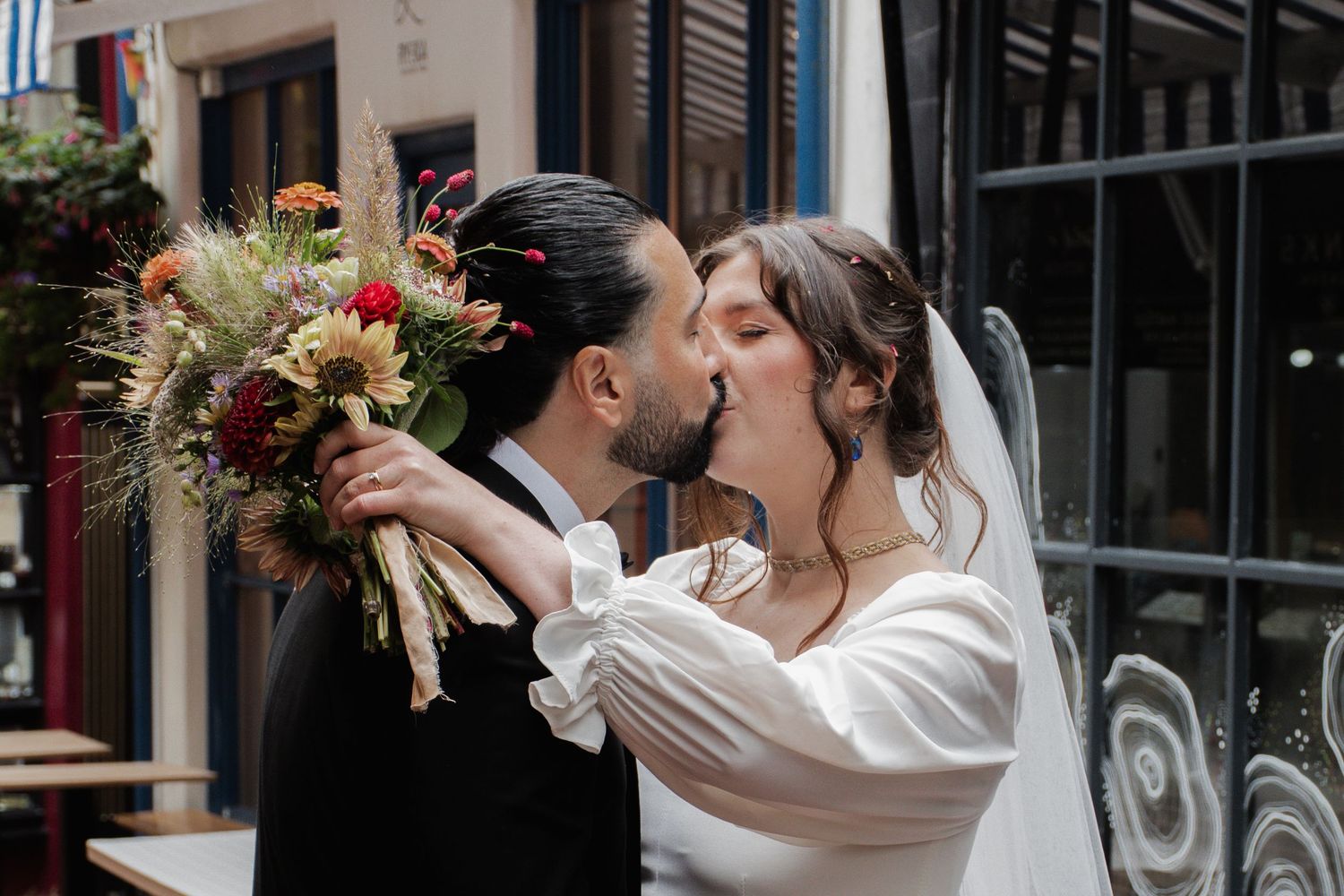 Bride and groom sharing a romantic kiss while holding a colorful autumn wedding bouquet outside a charming cafe.