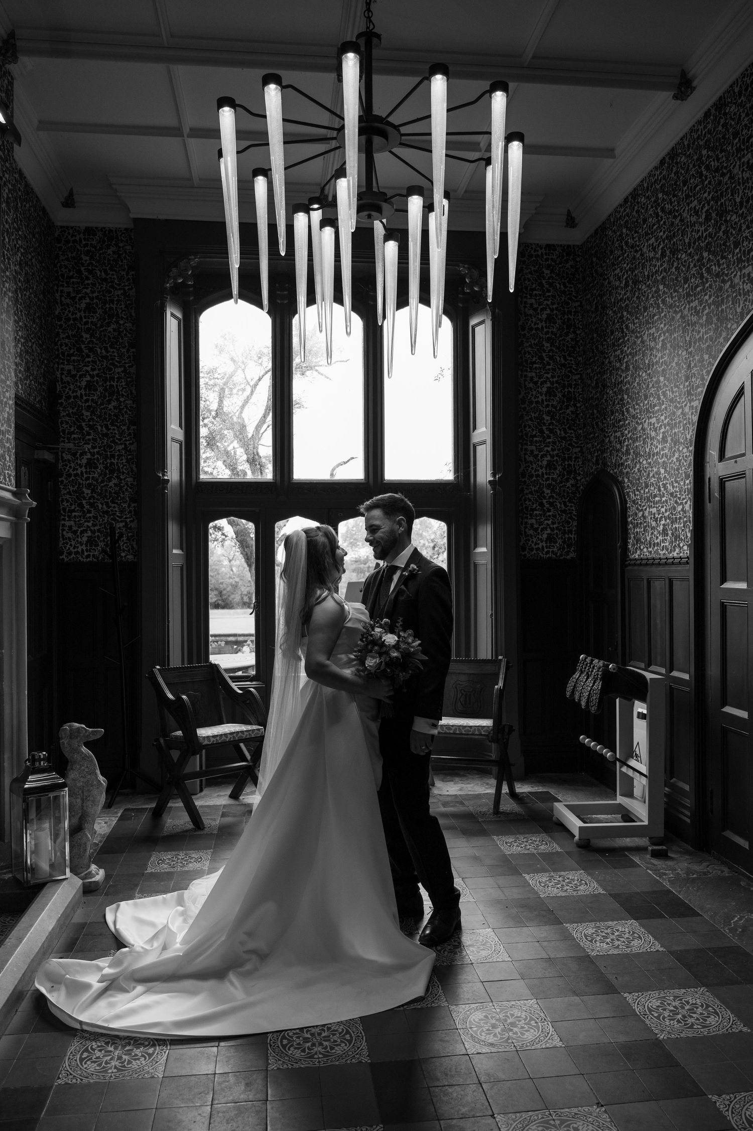 Elegant black and white photo of bride and groom embracing under a dramatic chandelier in a grand manor hall.