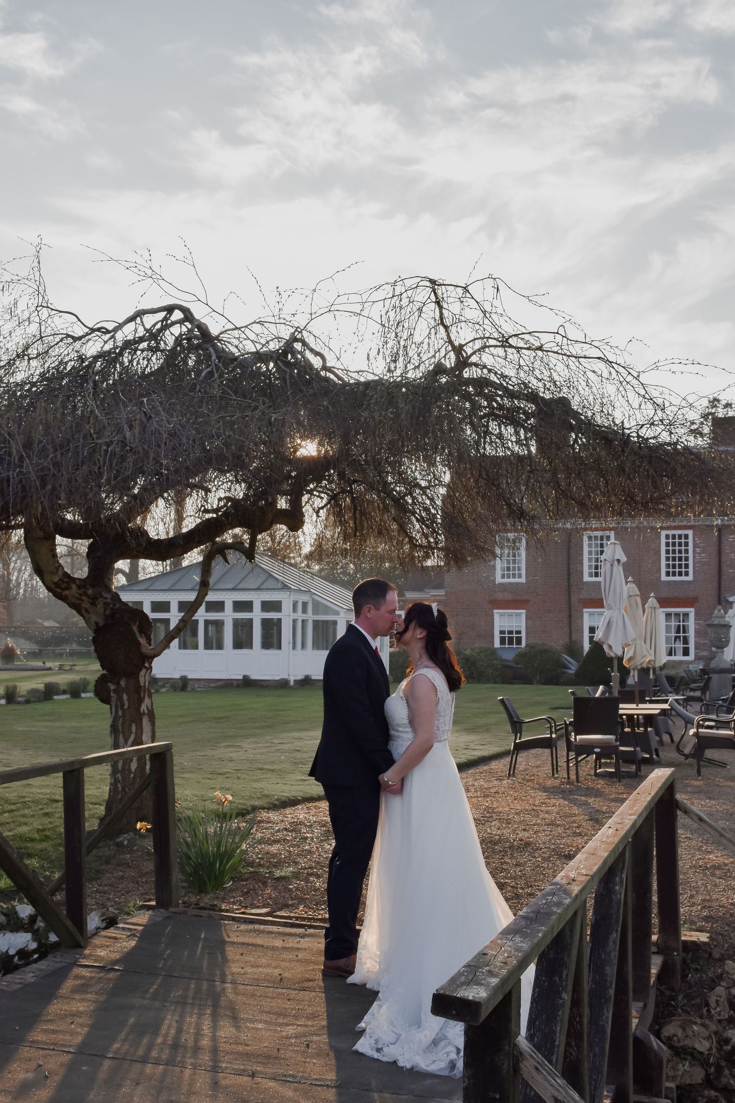Bride and groom kissing on a wooden bridge at sunset with a beautiful brick manor house in the background.
