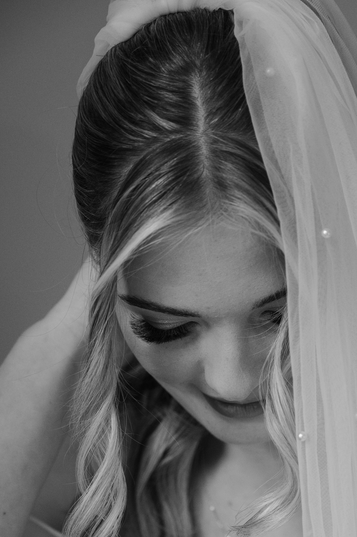 Black and white bridal portrait of blonde bride wearing veil with pearl details, looking down with dramatic eye makeup.
