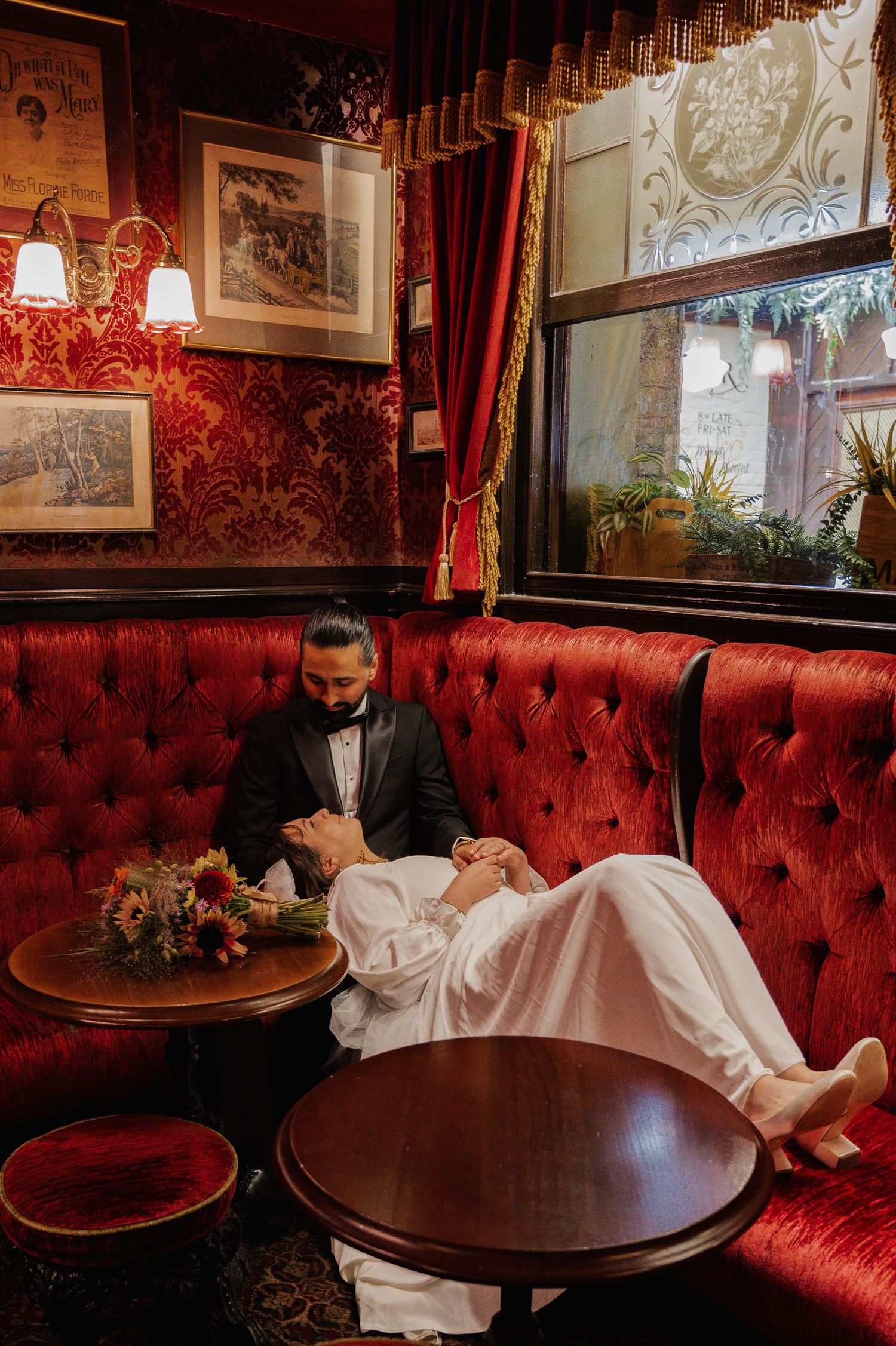 Bride reclines on a groom's lap in a cozy red velvet booth inside a warmly lit vintage pub after their wedding.