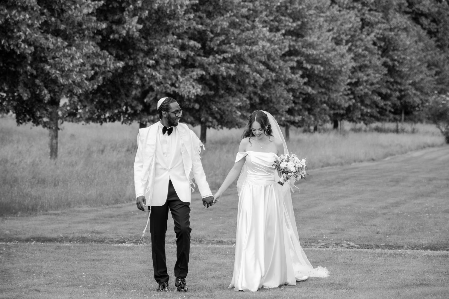 Black and white photo of a bride and groom holding hands walking through a lush green field on their wedding day.