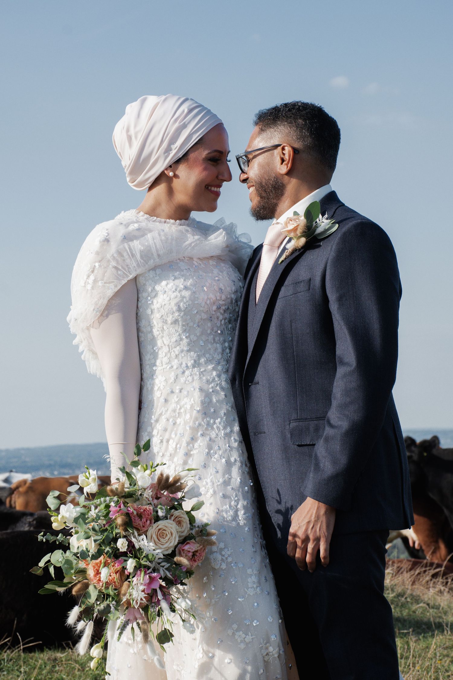Elegant bride in white hijab and embellished gown holds bouquet while gazing lovingly at groom in navy suit outdoors.