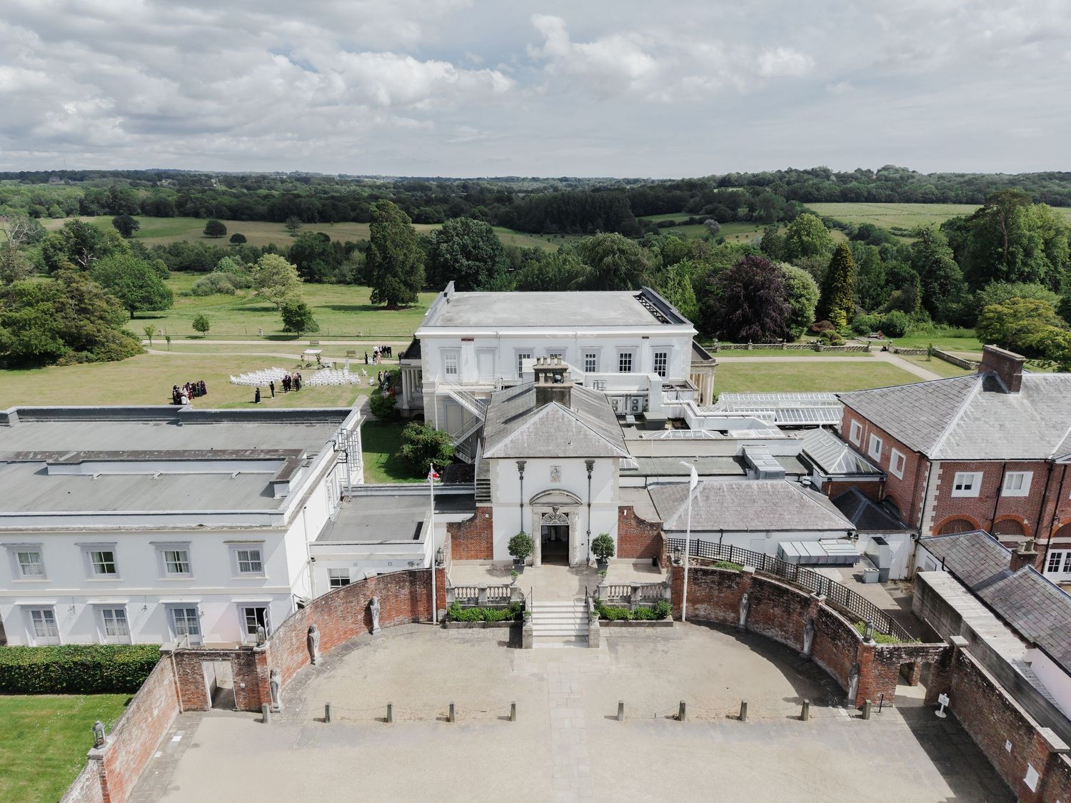 Aerial drone perspective of stately English country house with brick courtyard and scenic rural landscape.
