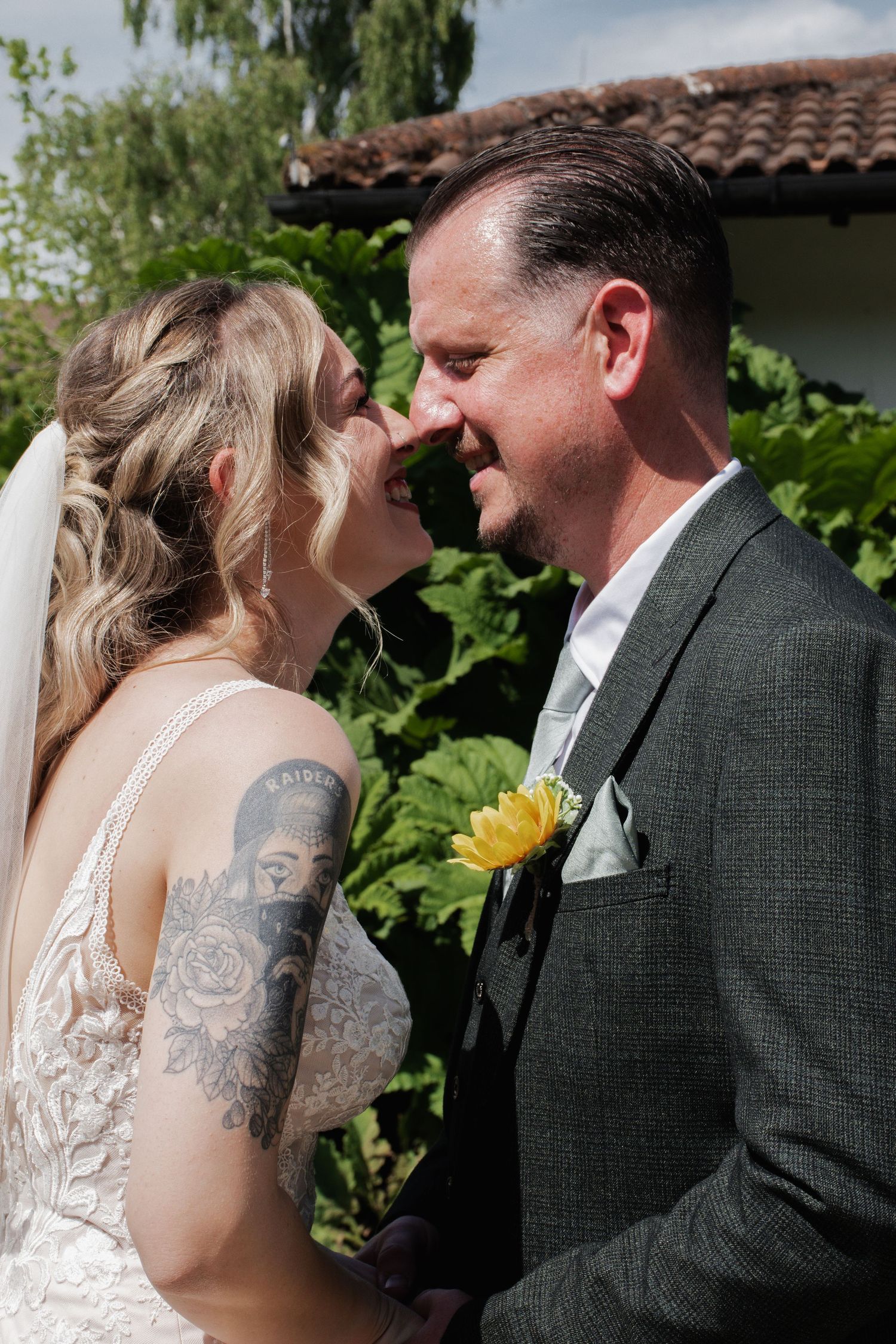 A tattooed bride and groom touch noses affectionately, groom wearing a gray suit with yellow boutonniere.