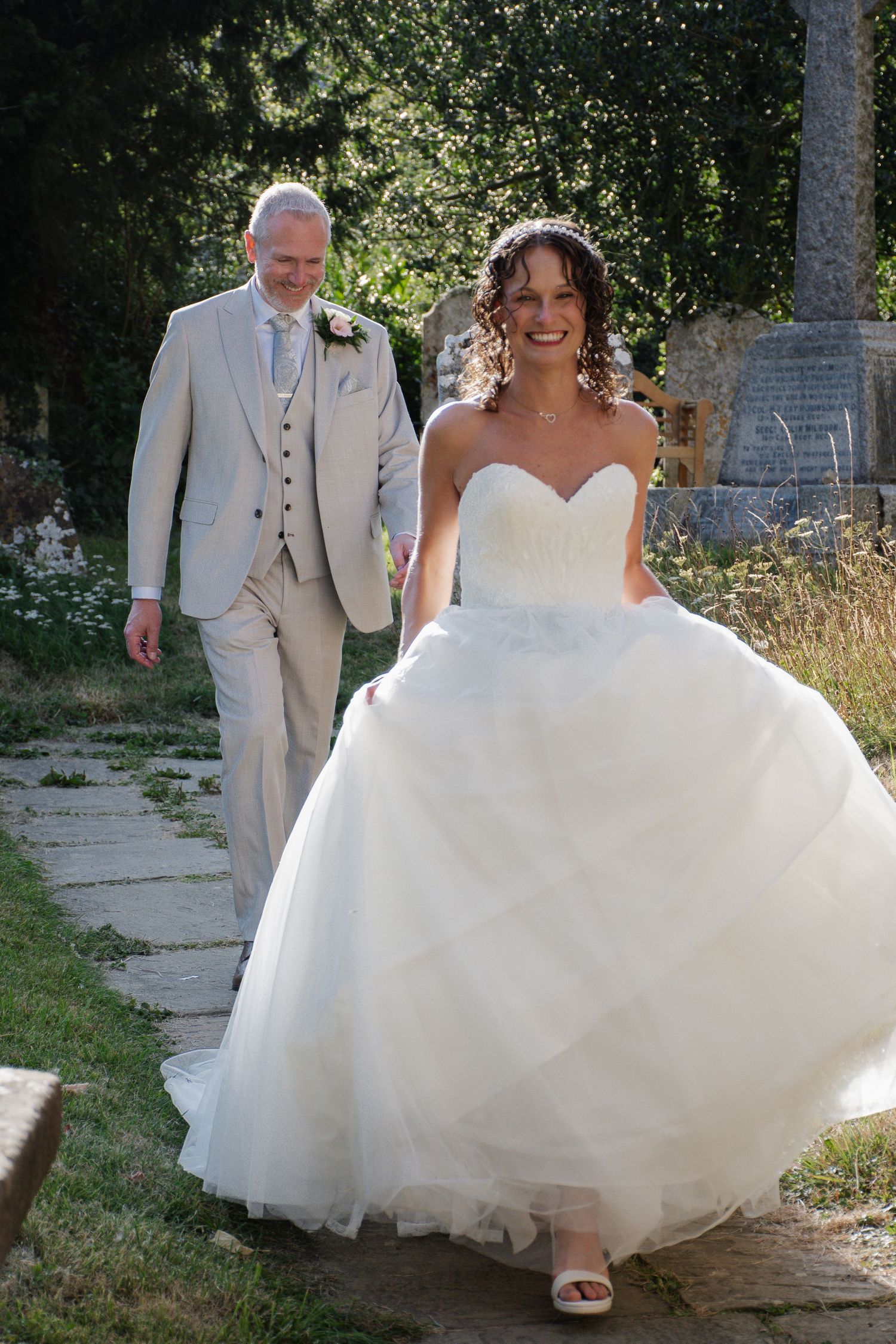 Radiant bride in strapless ballgown lifts skirt while walking garden path as groom in beige suit follows behind.