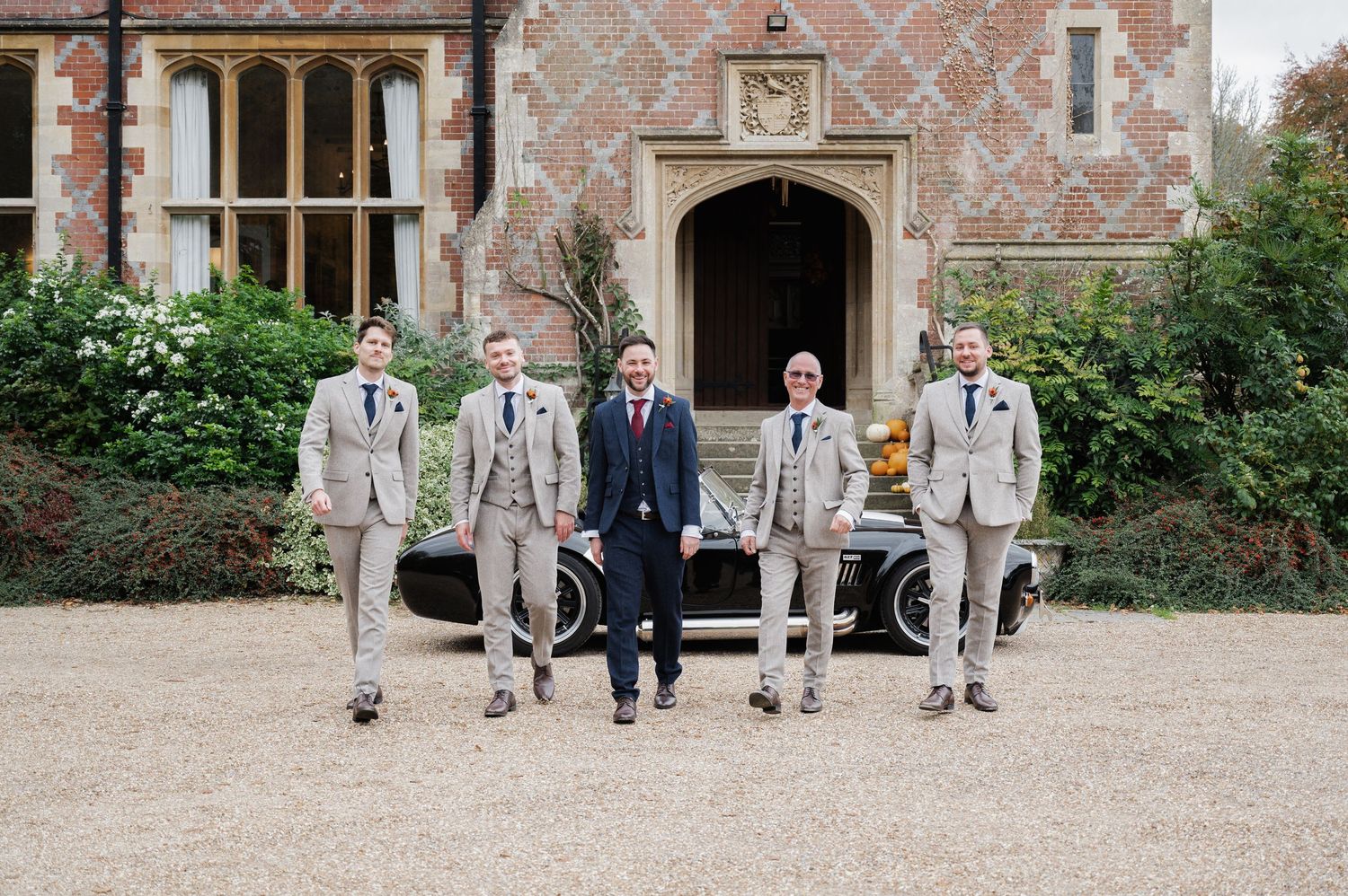 Groom and four groomsmen in suits posing confidently in front of a classic car at a manor house.