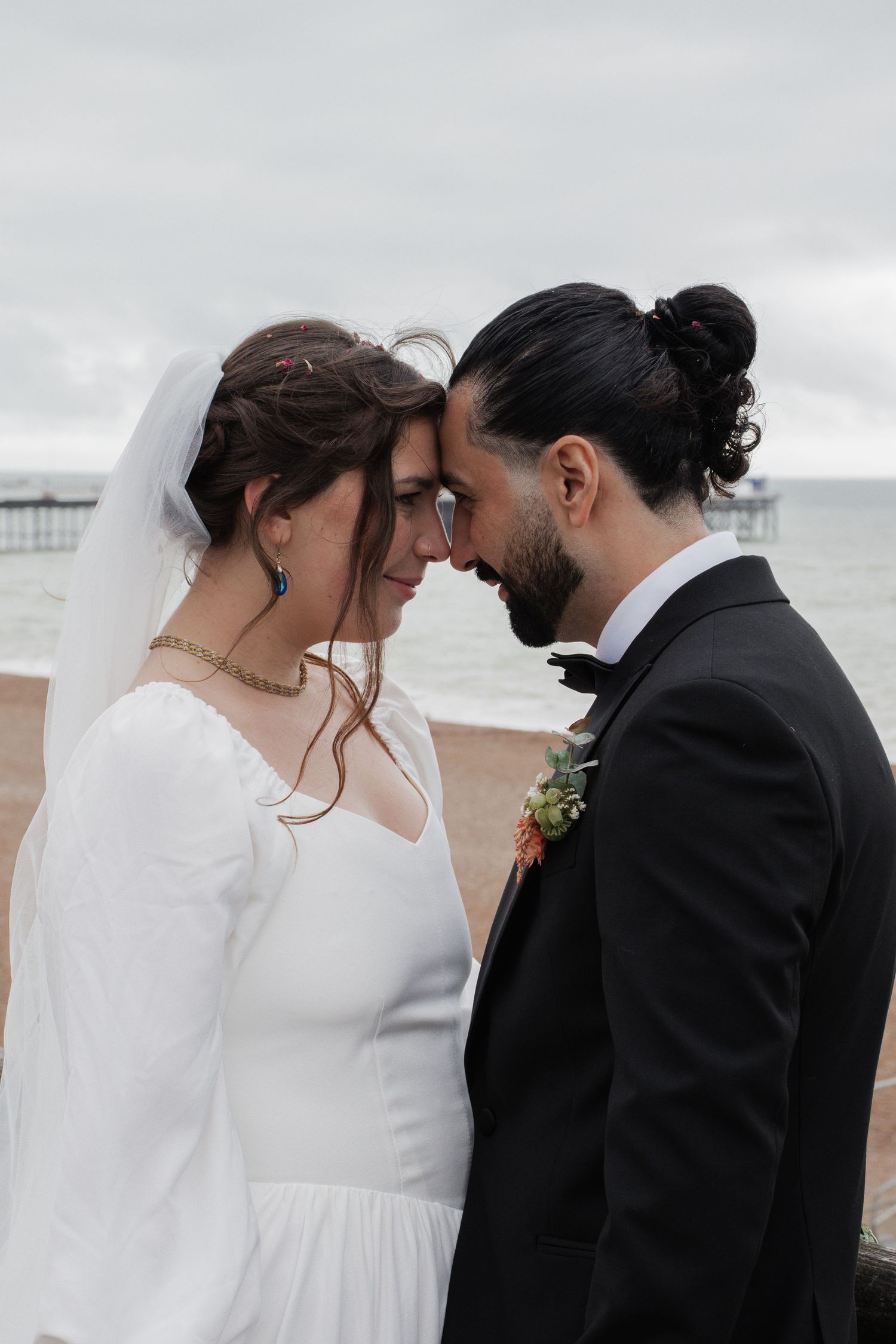 Bride and groom touch foreheads tenderly on a beach, with an overcast sky and pier visible in the background.