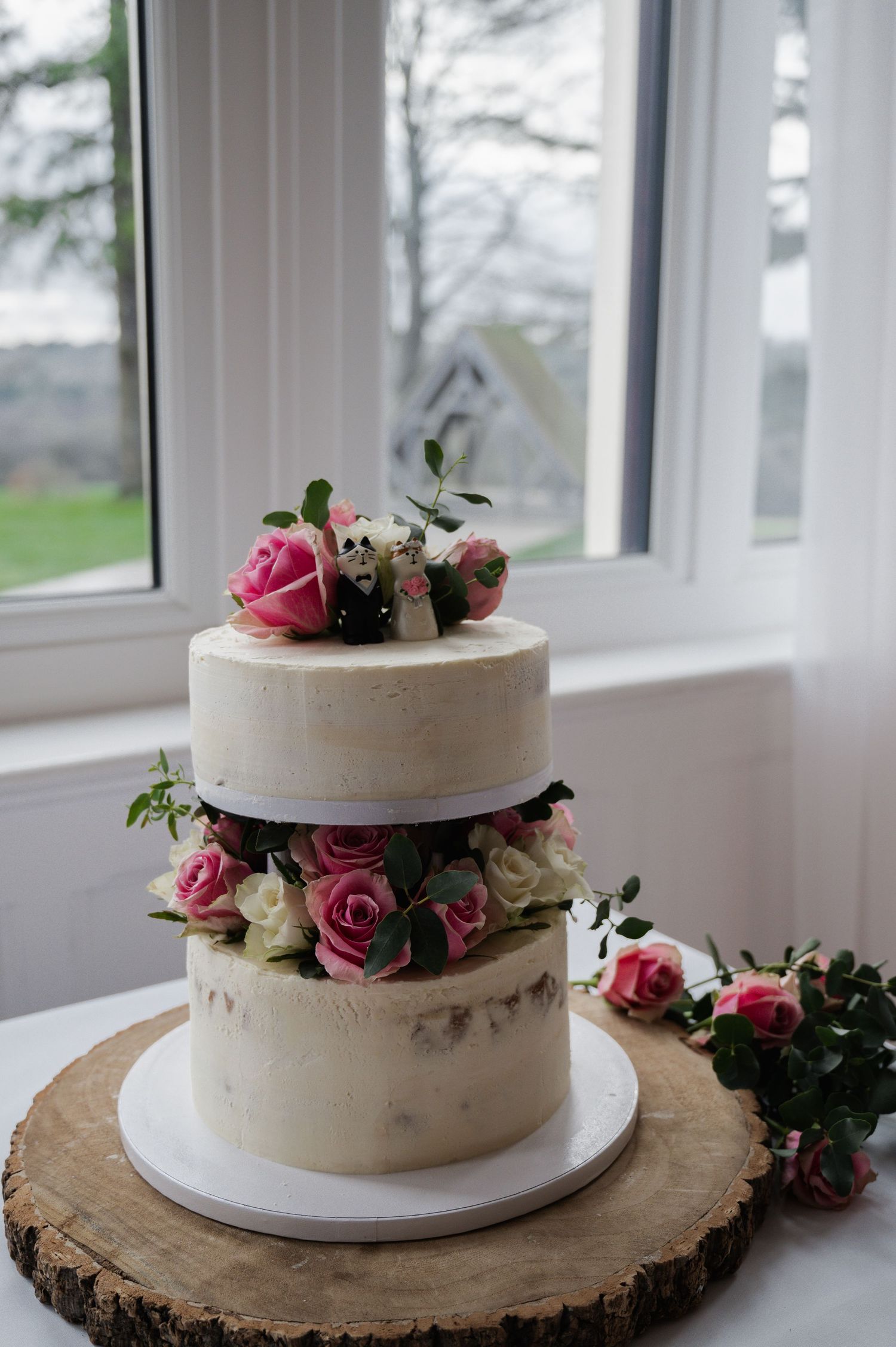 Two-tier semi-naked wedding cake decorated with pink roses on a rustic wooden stand.