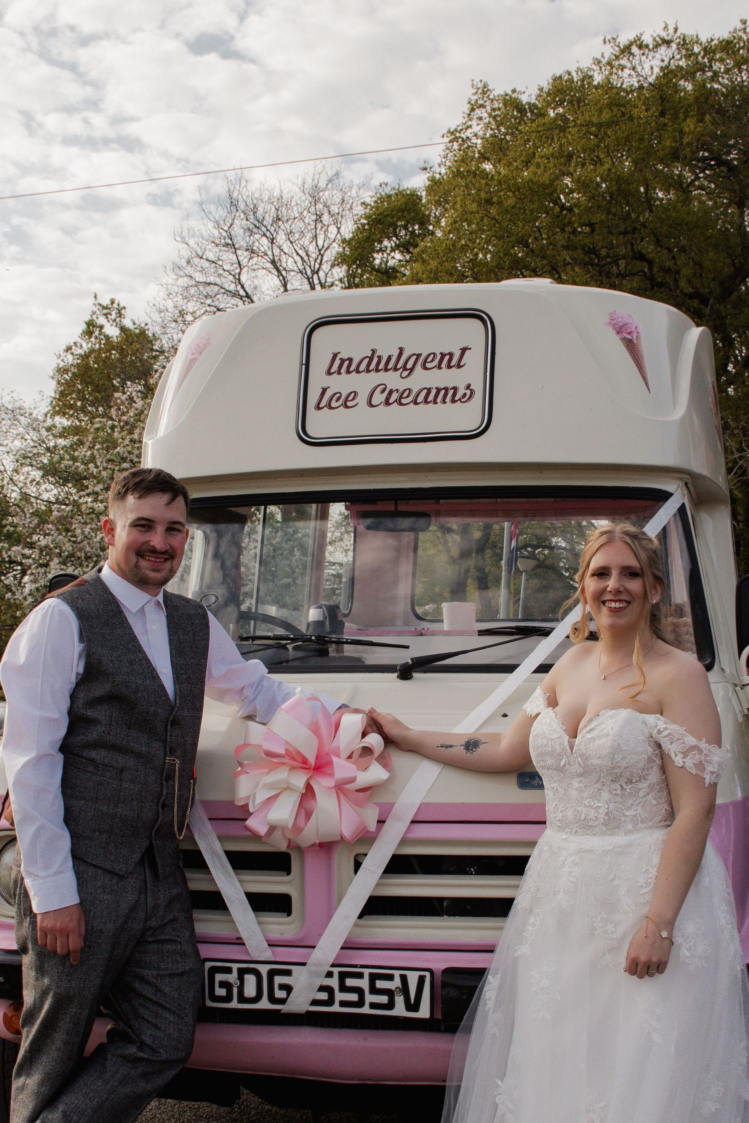 Bride and groom posing in front of a pink Indulgent Ice Creams van decorated with a pink bow on their wedding day.