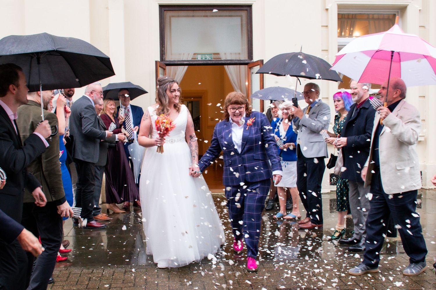 A joyful same-sex couple exits a building through a shower of colorful confetti as guests celebrate with umbrellas.