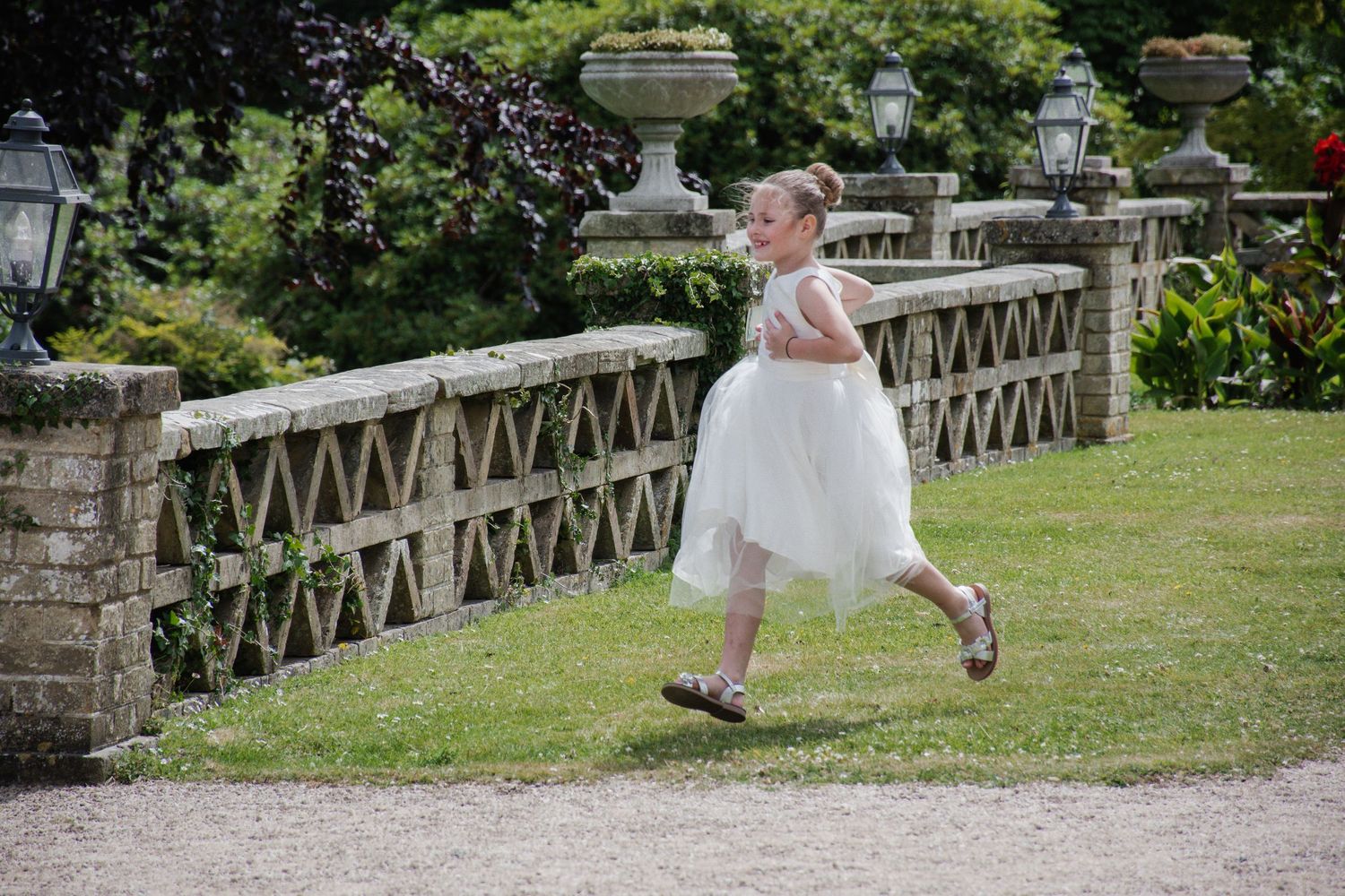 A young flower girl in a white tulle dress runs along a stone garden path.