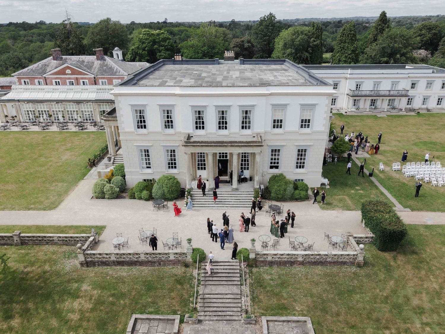 Aerial view of a grand white Georgian manor house with wedding guests gathered on the steps and grounds. Buxted Park Hotel