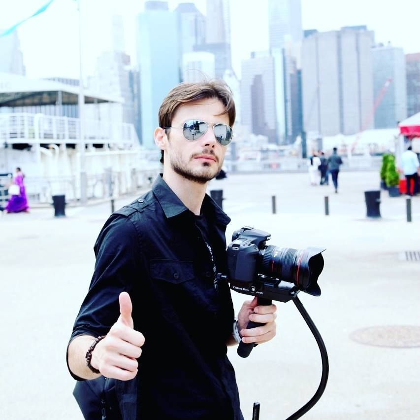 A photographer wearing dark clothing and sunglasses holds a professional camera while shooting outdoors in an urban setting.
