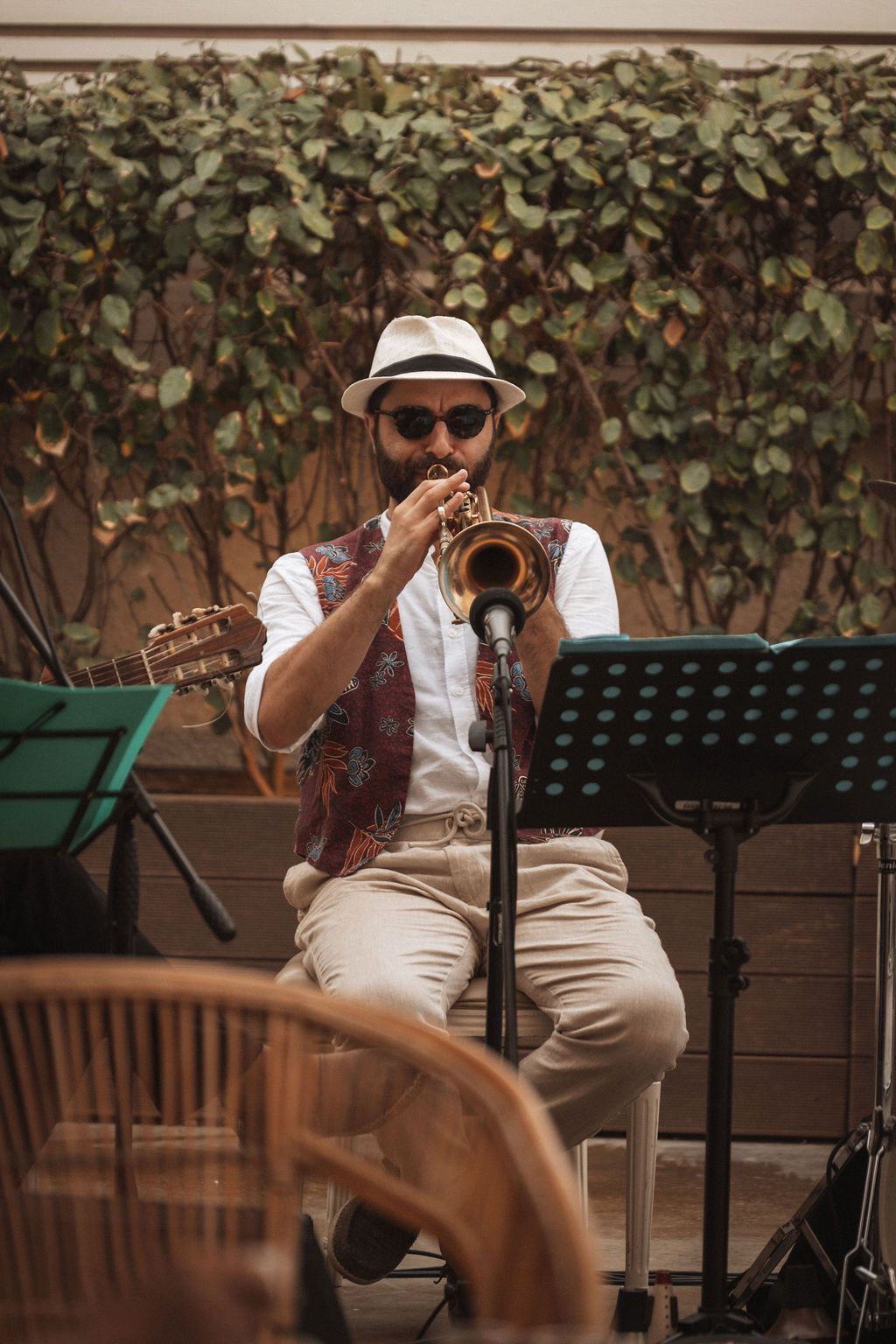 Musician in a fedora and vest plays trumpet against an ivy-covered wall backdrop.