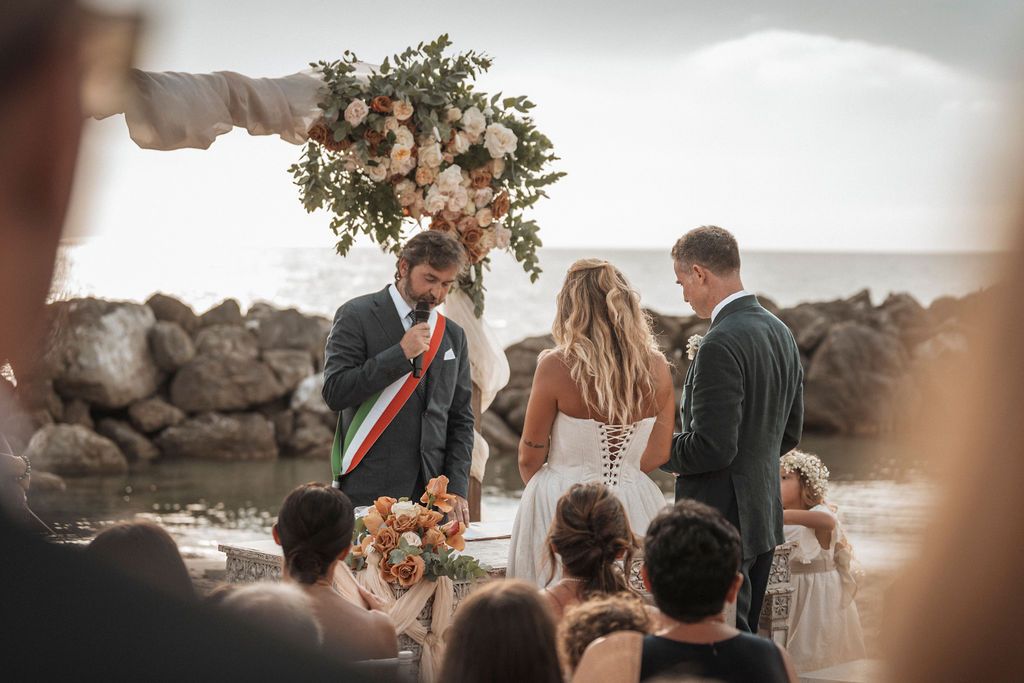 Wedding ceremony setup on a beach deck with string lights and floral arrangements at sunset.
