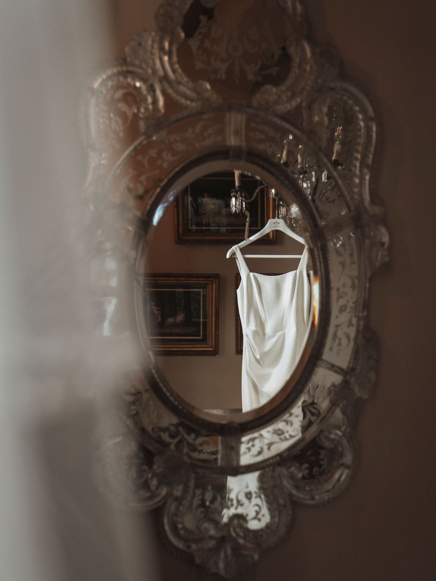 White wedding dress hanging in an ornate vintage silver mirror reflecting a chandelier and elegant room decor.