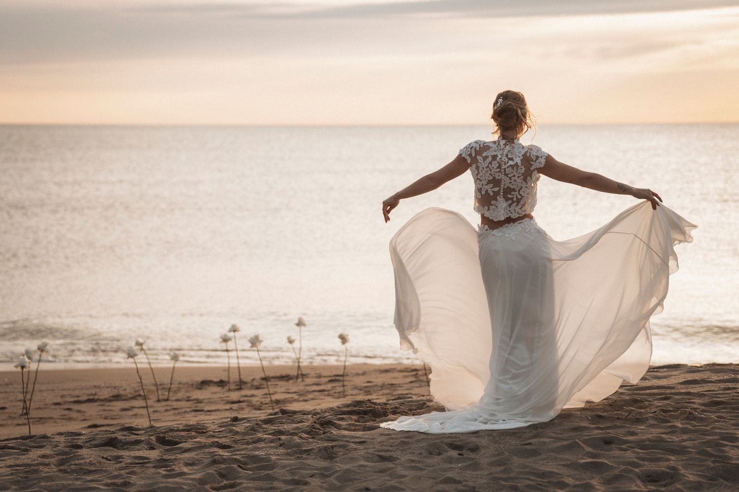 Flowing white wedding dress captured against ocean sunset backdrop on sandy beach.