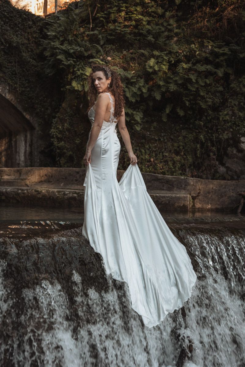 A white dress flows gracefully against a cascading waterfall backdrop in a forest setting.