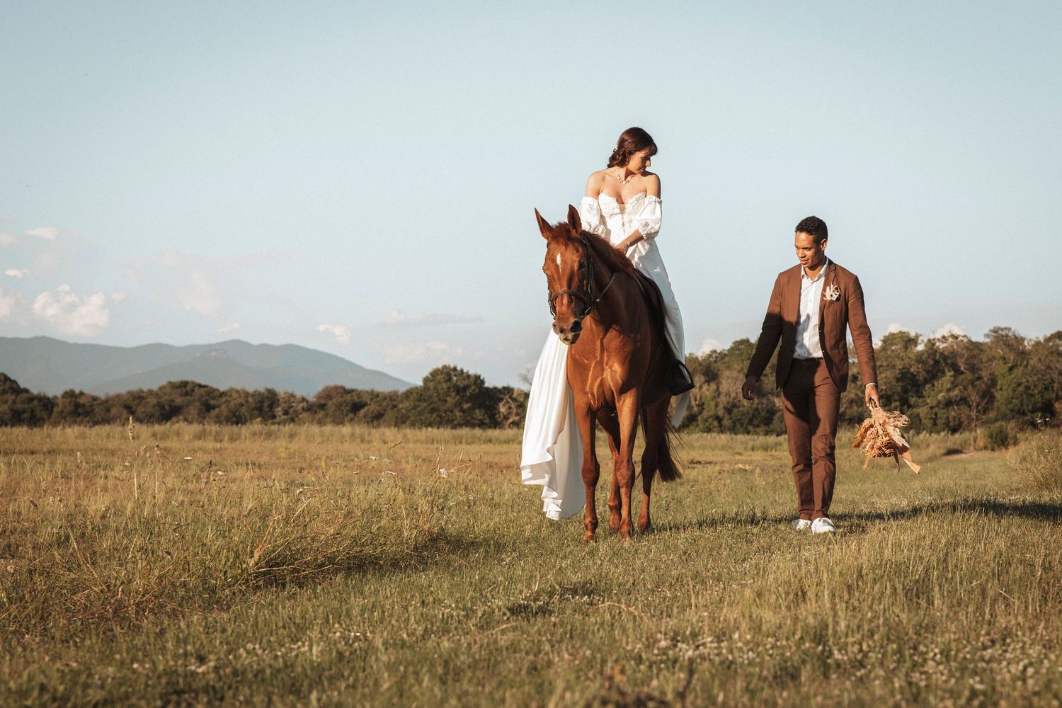 Wedding couple posing with horses in a scenic meadow landscape with mountains in background.