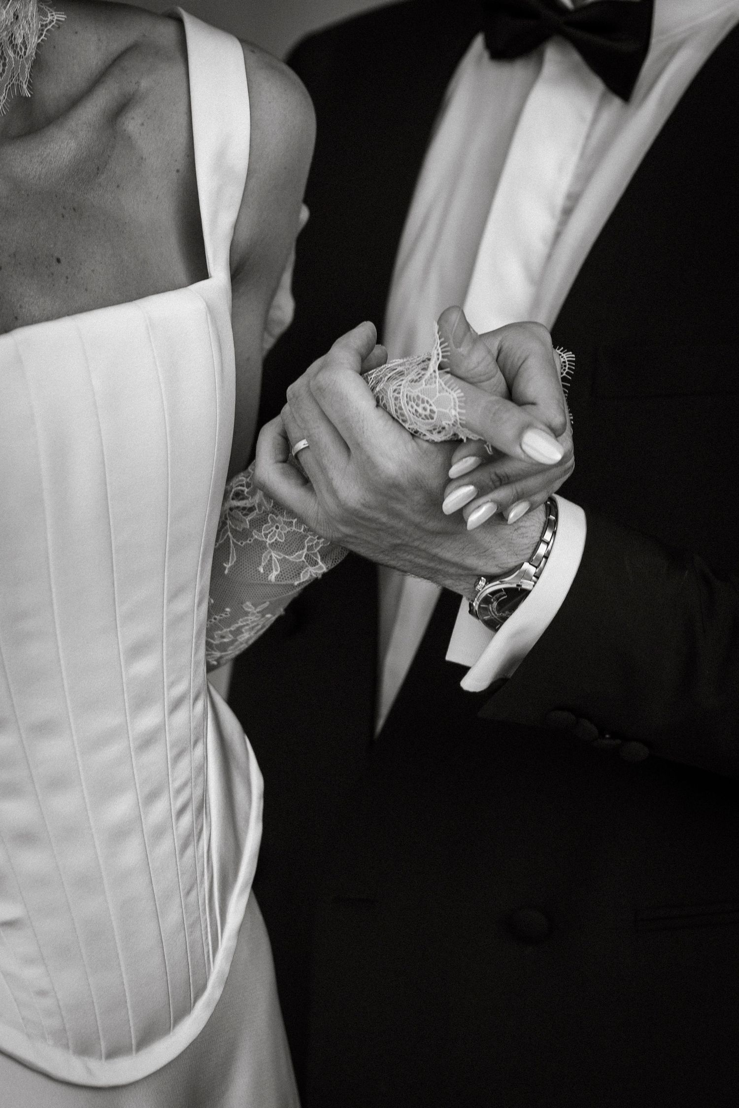 Black and white photo of bride and groom holding hands, showing wedding attire details including lace dress and tuxedo.