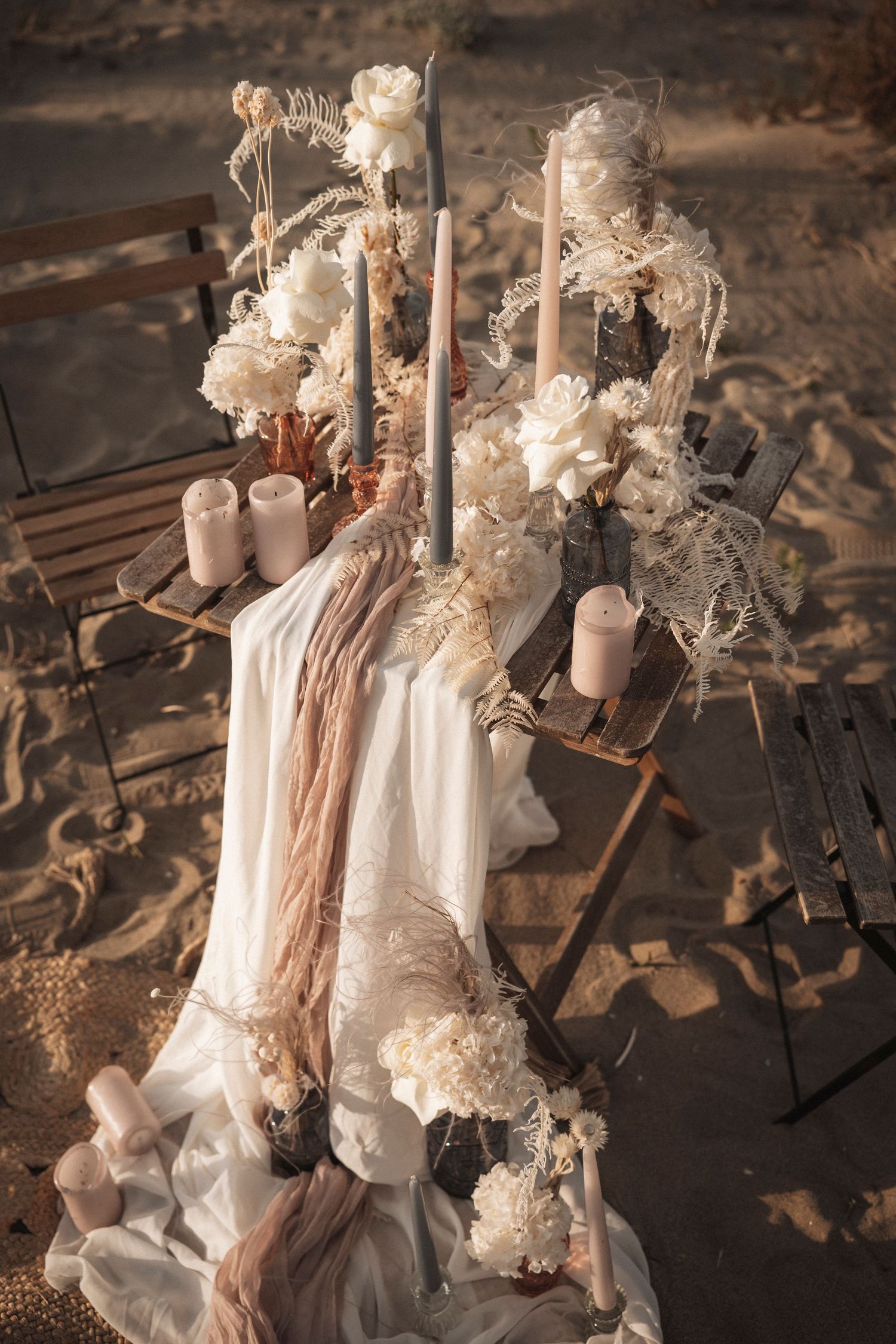 Rustic wedding decor featuring white candles, dried flowers, and flowing fabric on a weathered wooden surface.