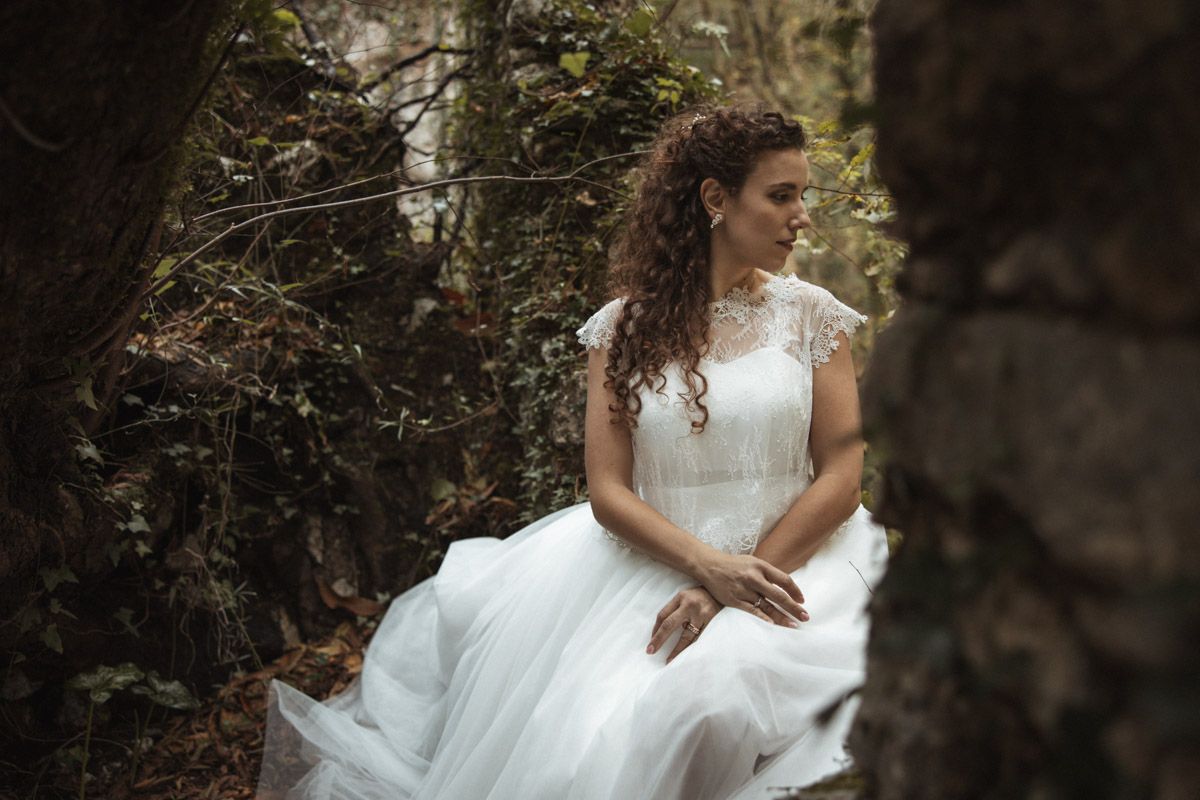 A bride in a white wedding dress poses in a moody forest setting with dramatic lighting and shadows.
