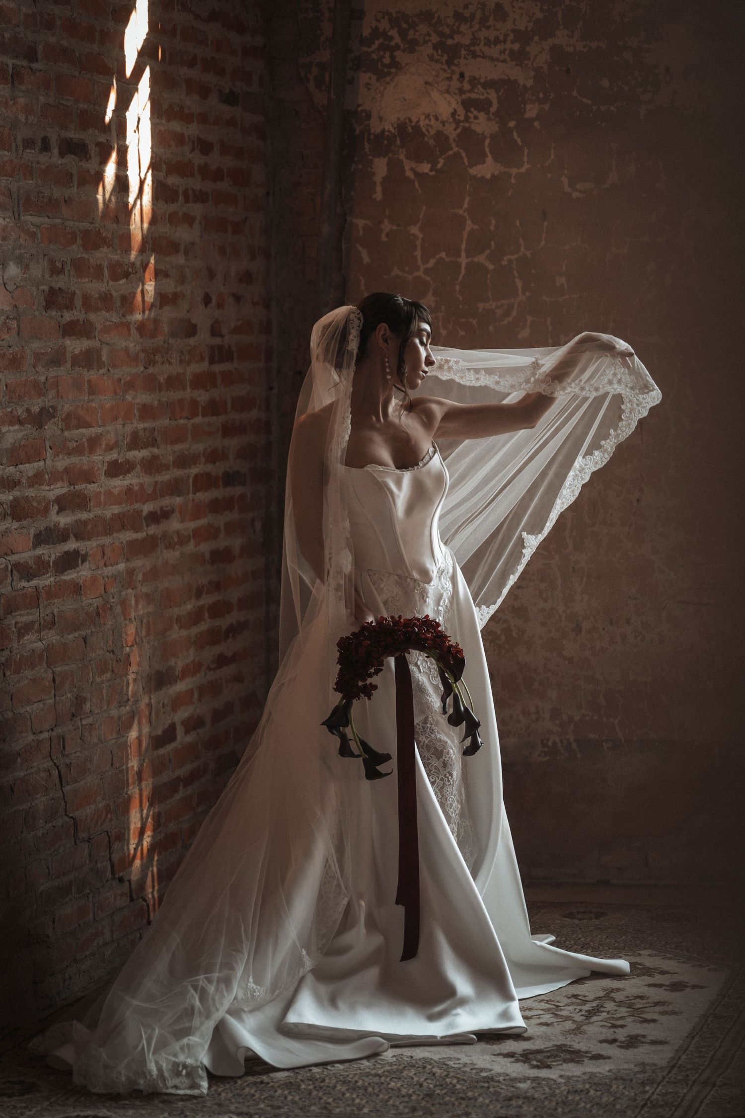 Dramatic side view of wedding dress and veil in brick room with streaming sunlight.