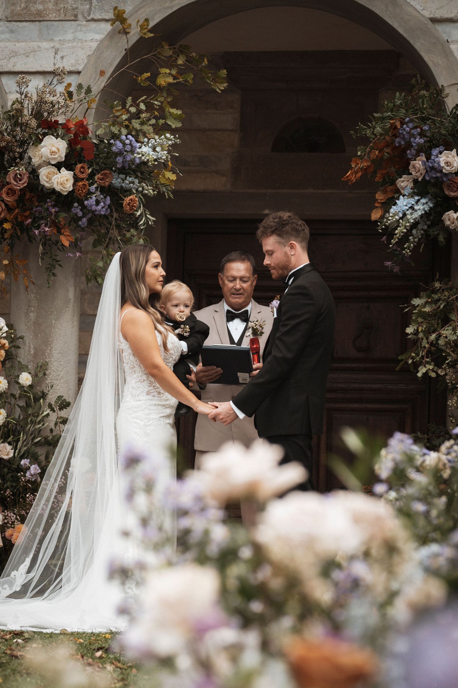Wedding ceremony under stone archway decorated with floral arrangements and greenery.
