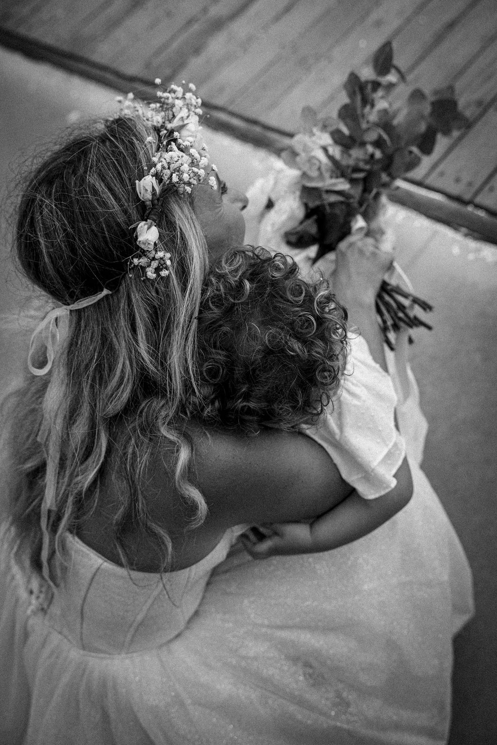 Black and white photo of a bride with flower crown embracing a small child.