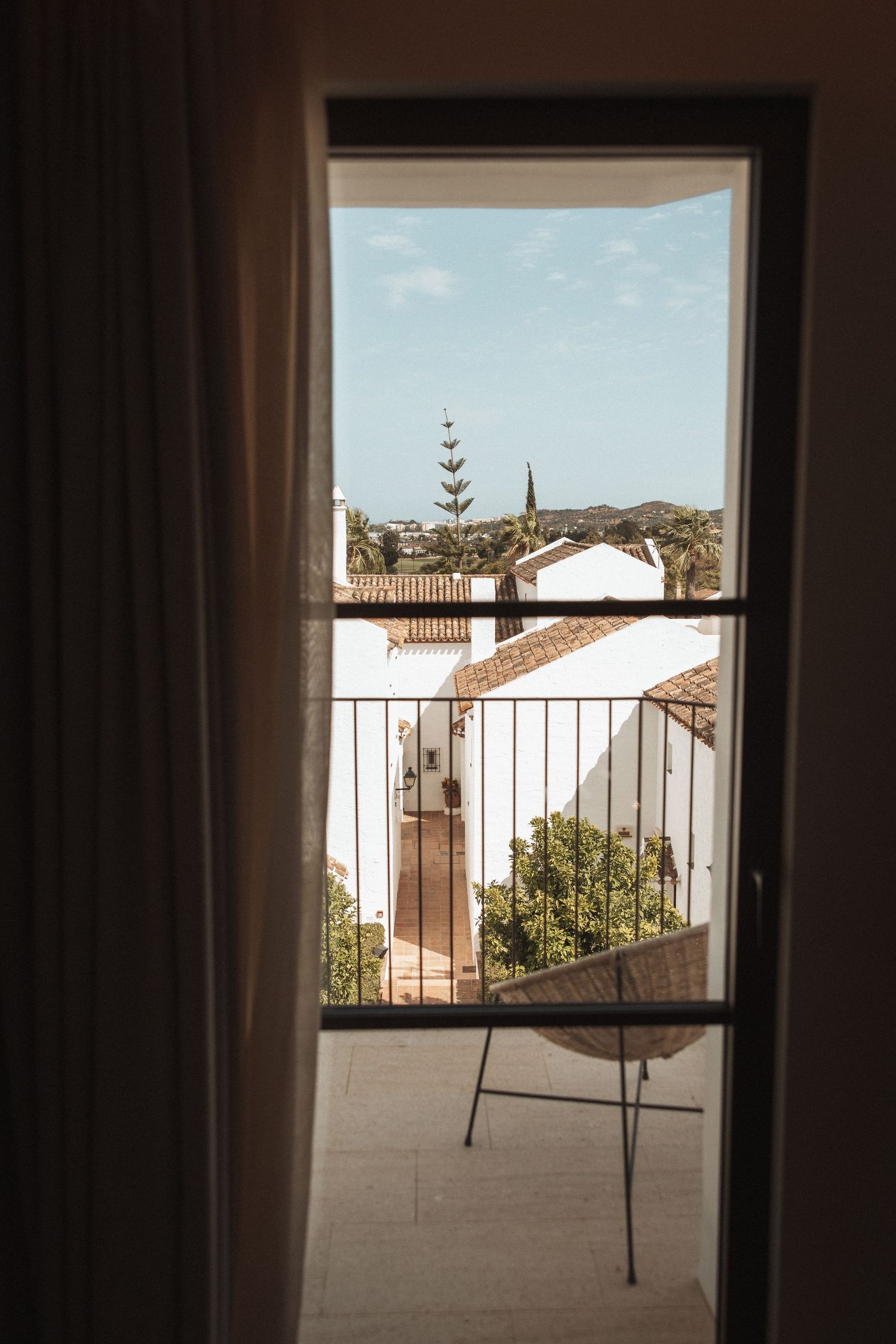 Sunlit balcony view through curtained windows overlooking Mediterranean-style rooftops.
